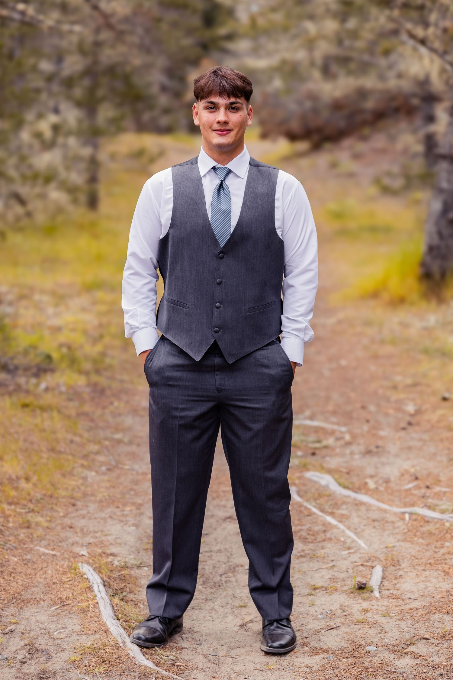 Young man posing outdoors in a gray vest, dress pants, and blue tie for a prom photo session.