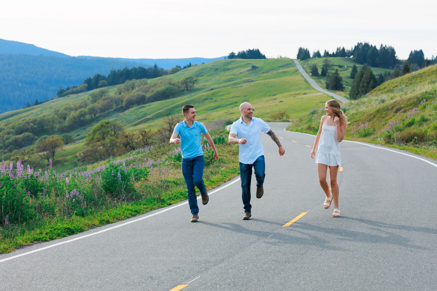 Family of three running and laughing on scenic mountain road with green hills behind.