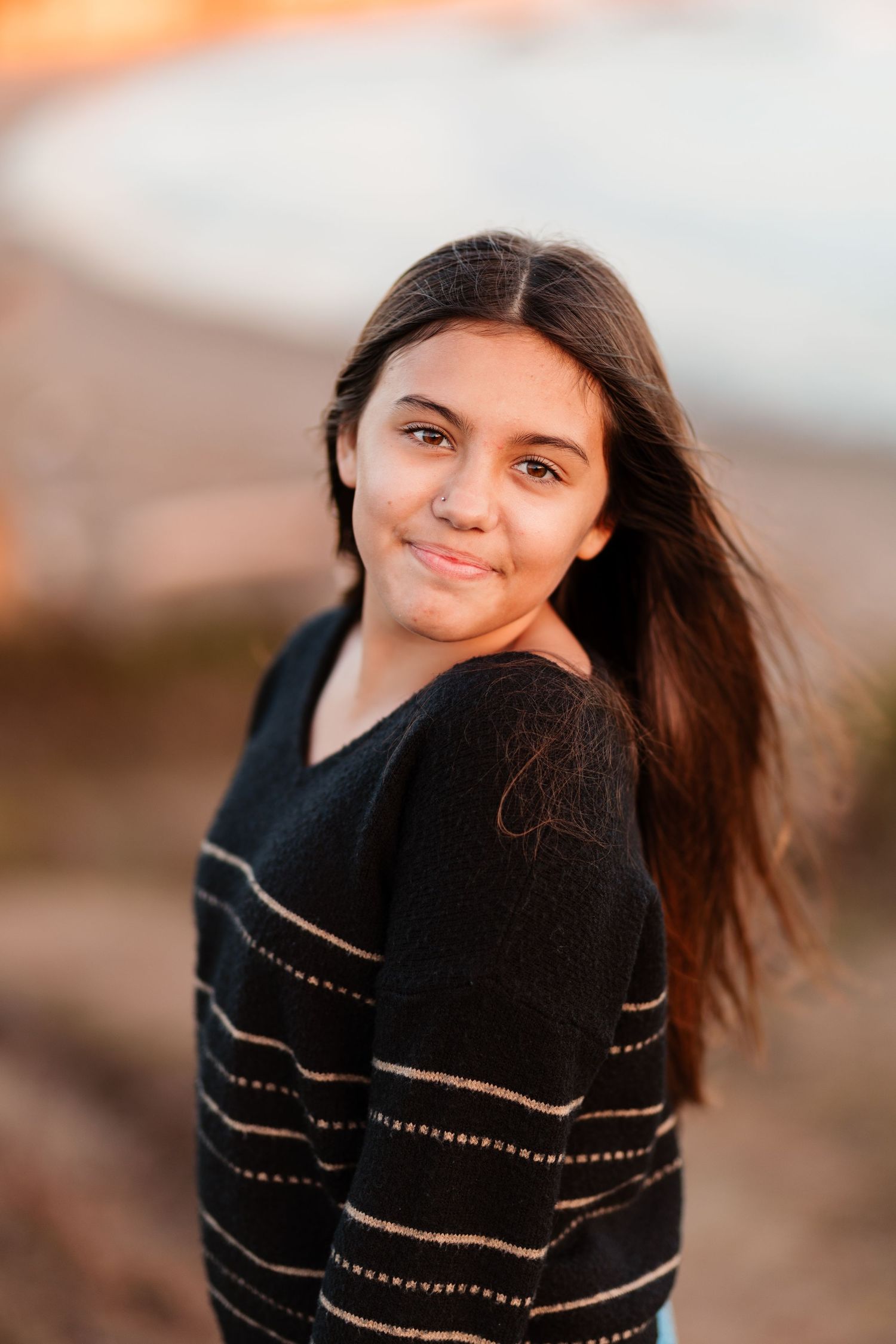 Young teenage girl with long dark hair smiling over shoulder wearing black striped sweater at sunset beach.