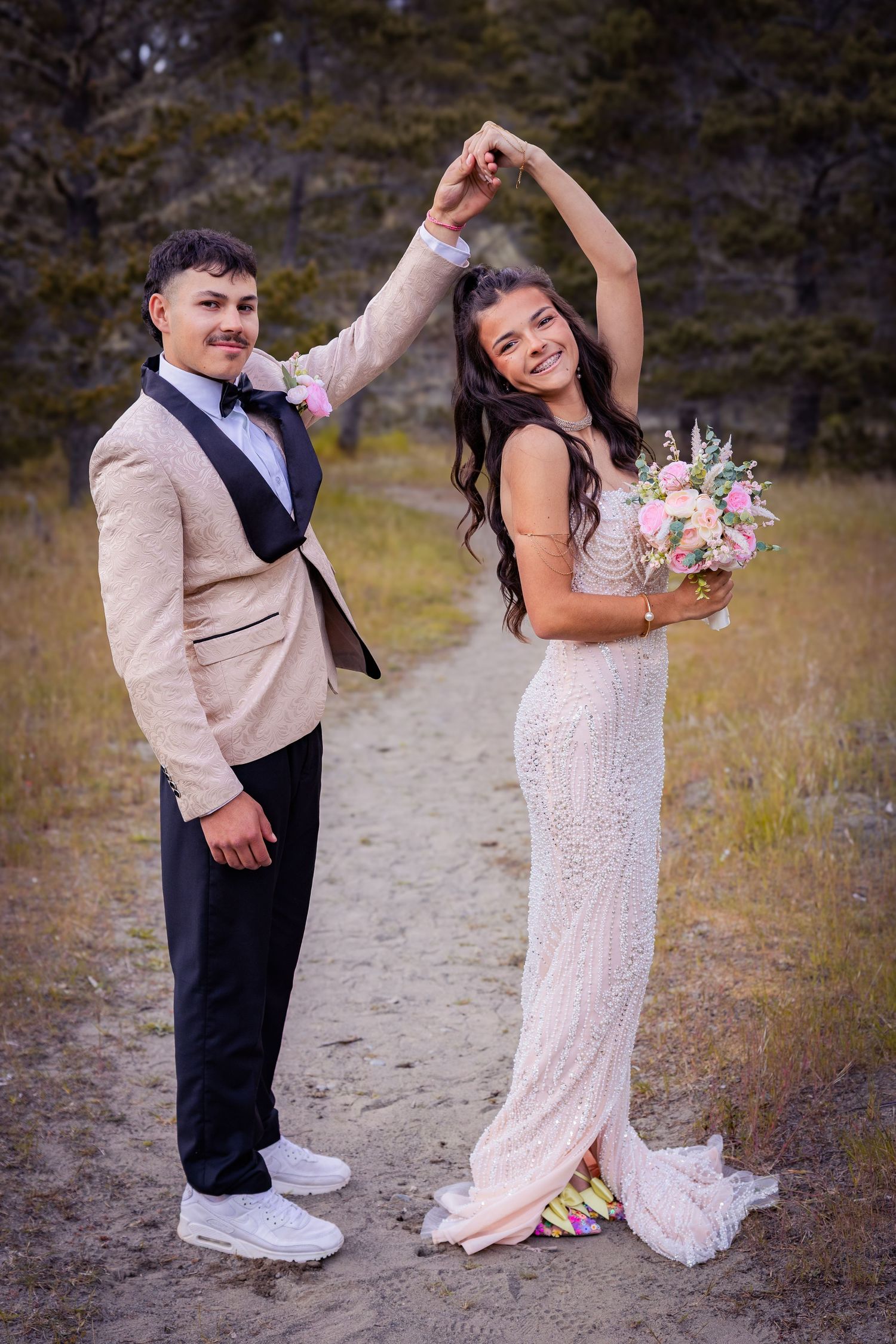 Bride and groom dancing outdoors, she holds bouquet in white gown, he wears beige tuxedo on forest path.