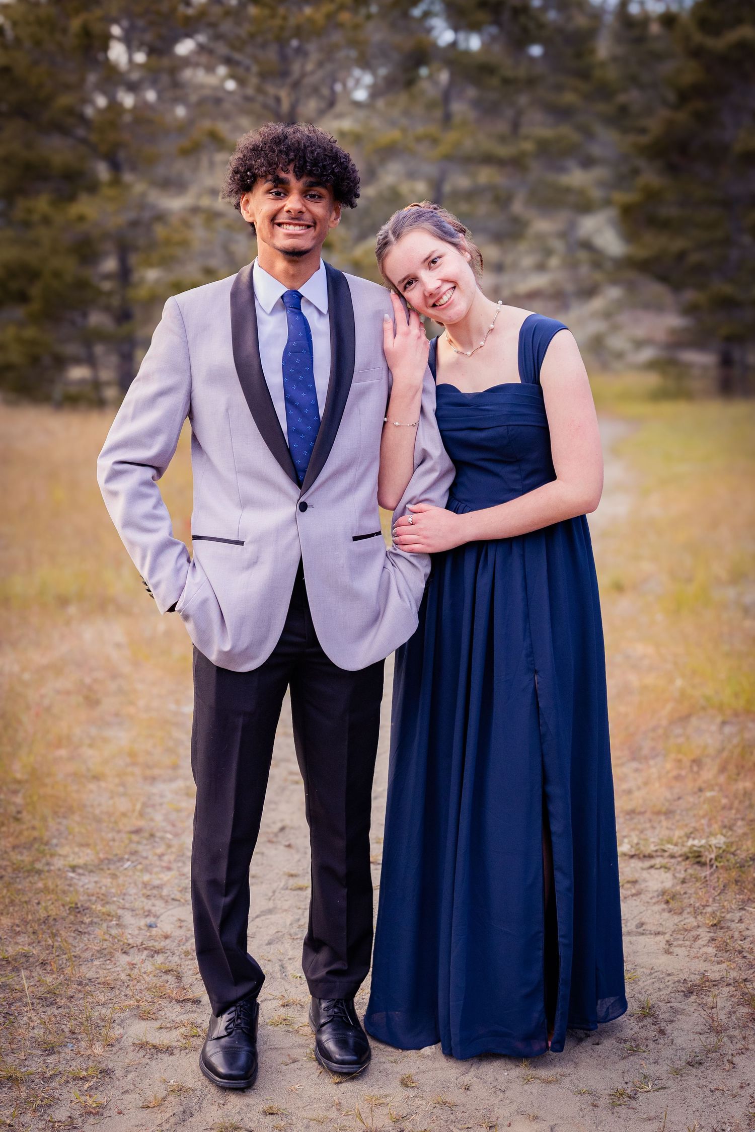 A young couple dressed in prom attire, man in gray tuxedo and woman in navy gown, posing outdoors.