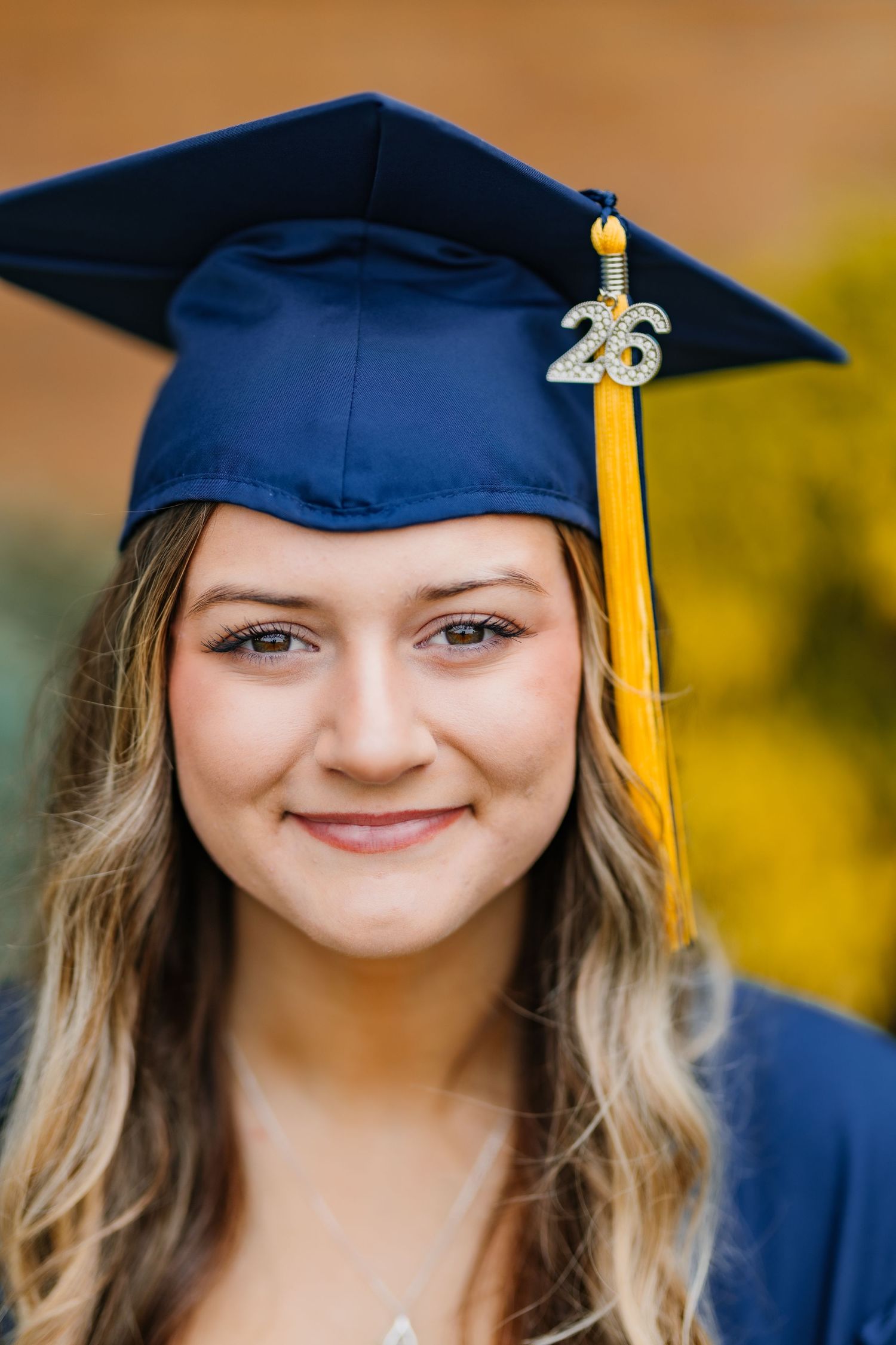 Close-up portrait of smiling senior girl wearing navy graduation cap with gold tassel and Class of 26 charm.