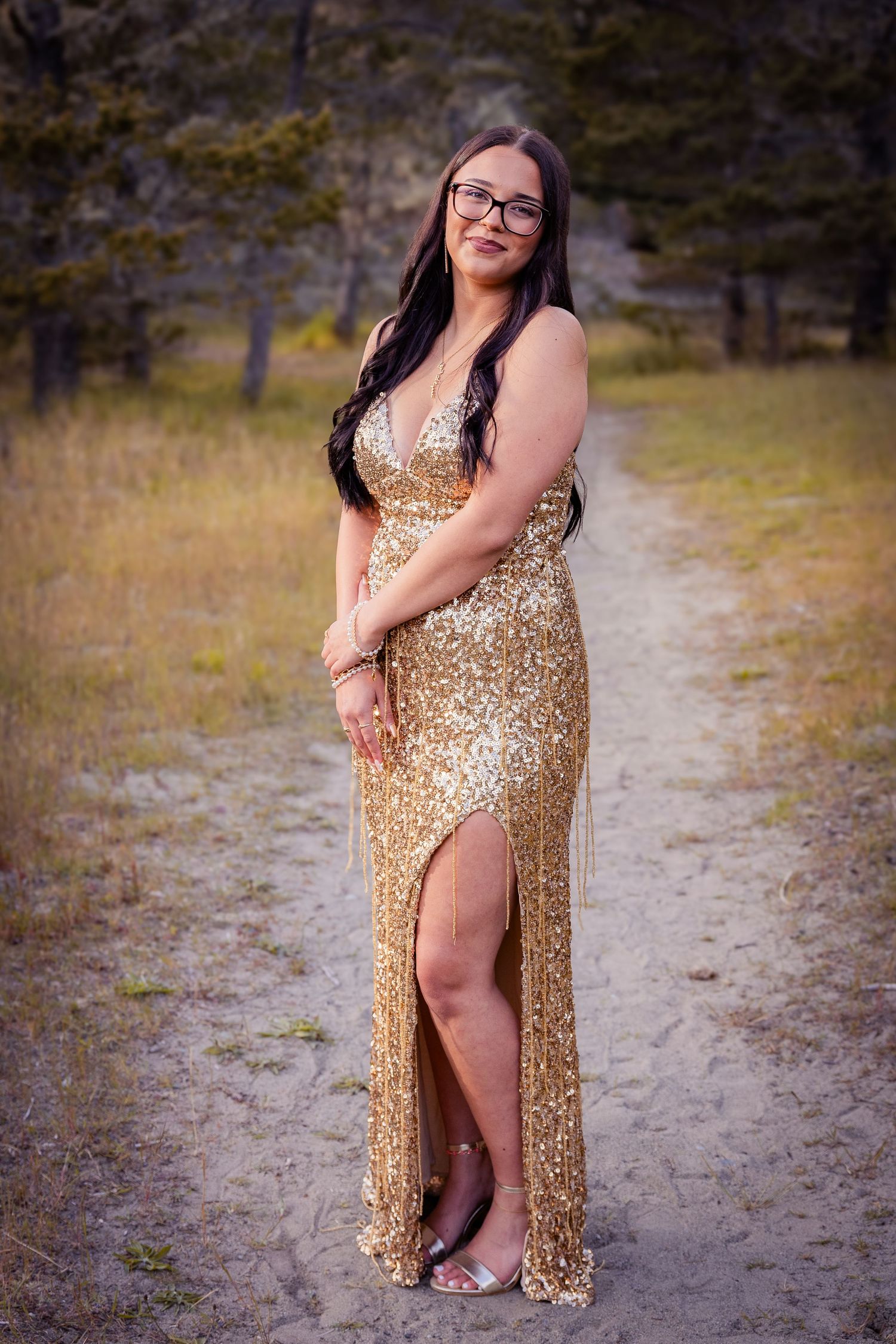 Woman in gold sequin gown with slit poses on sandy path surrounded by trees during golden hour.