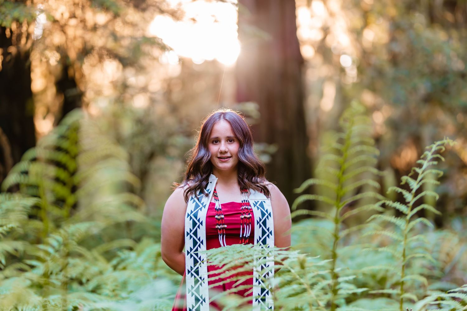 Woman in colorful dress standing among ferns in sunlit redwood forest.