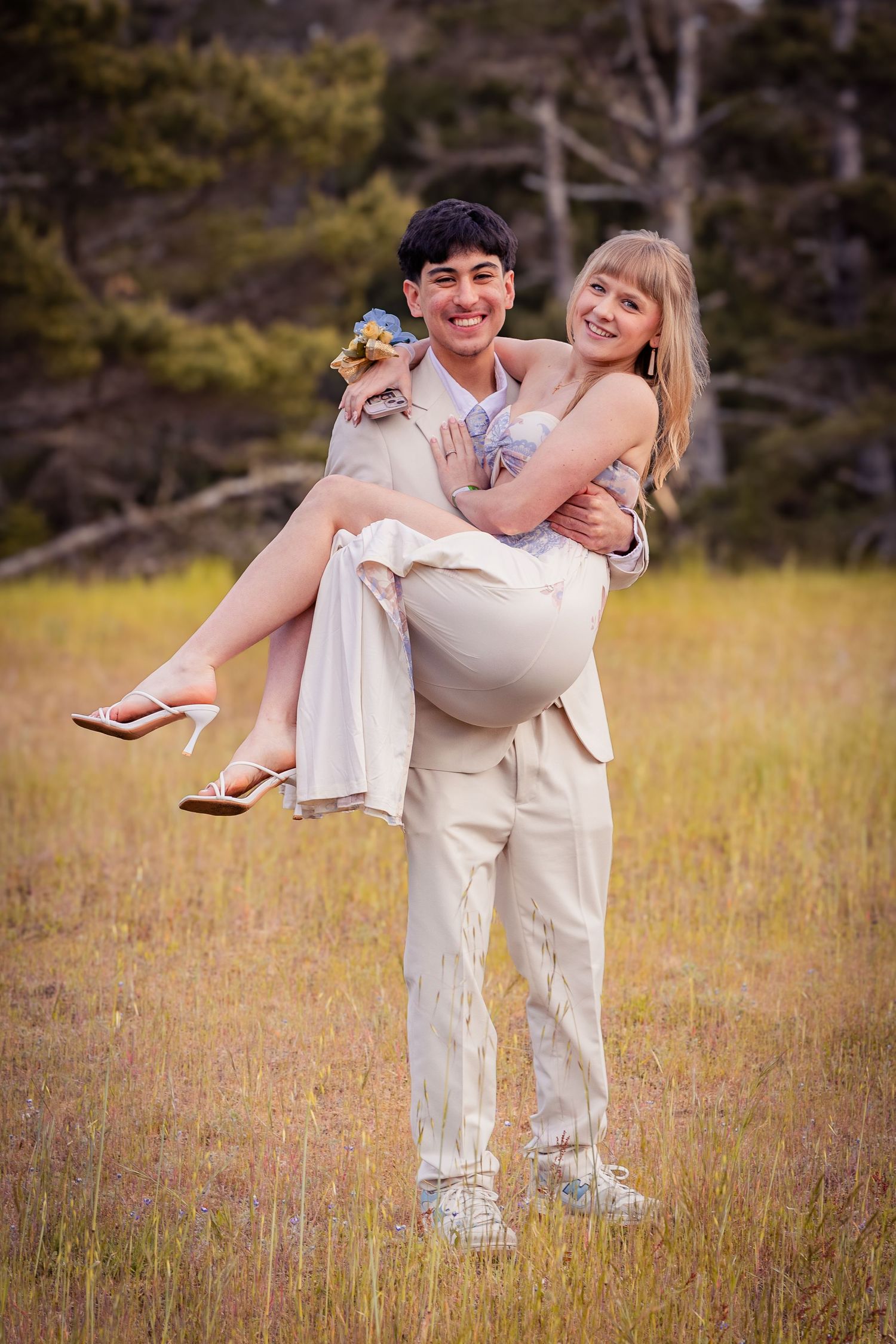 Young couple in formal attire posing in a meadow, man carrying woman in his arms, both smiling outdoors.