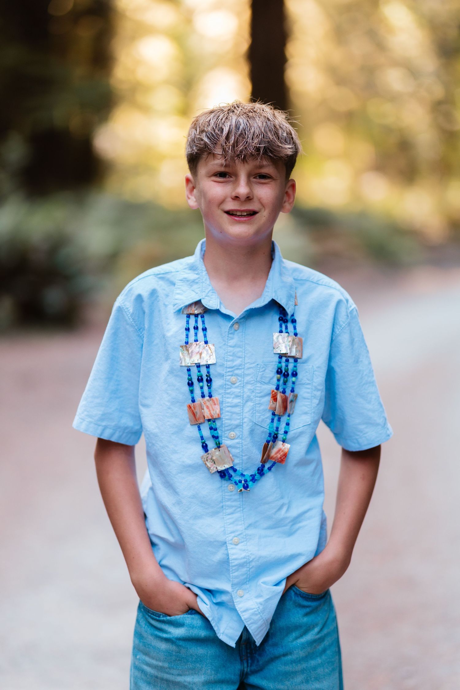 Smiling boy in blue shirt wearing decorative Native American beaded necklace, standing outdoors in a forest setting.