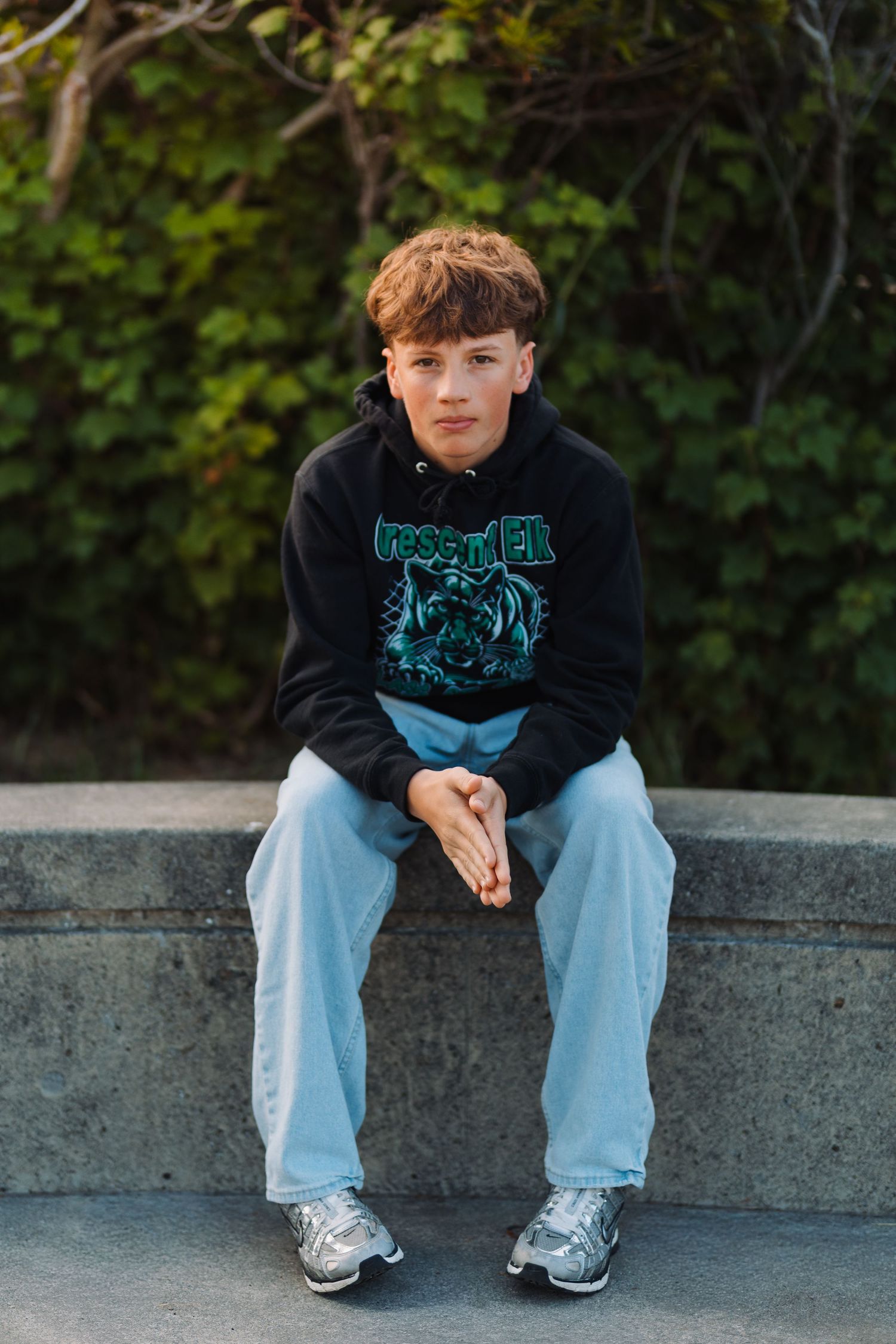 Teen boy in black graphic hoodie and light jeans sits on stone ledge outdoors with green foliage background.