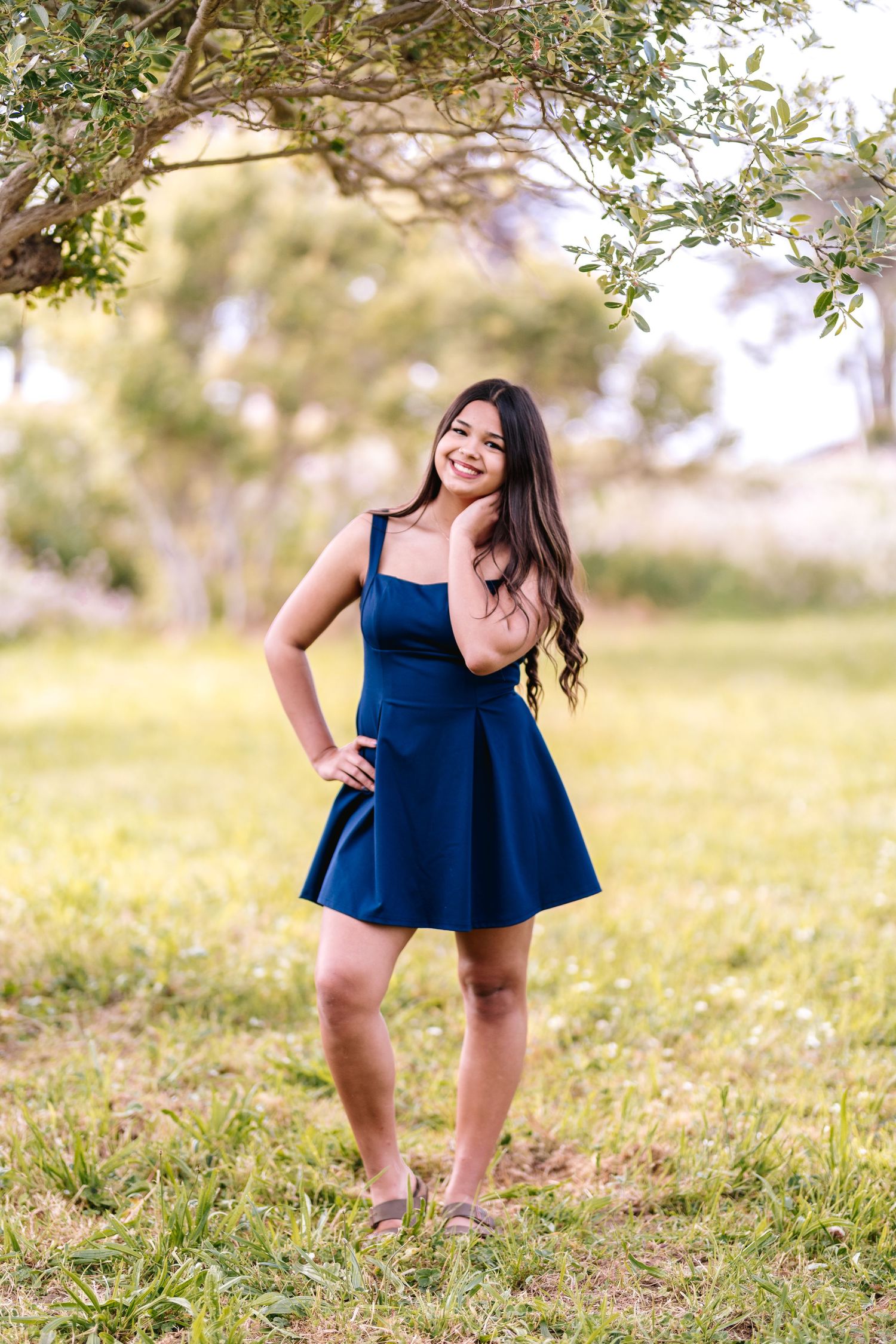 Young woman in navy blue skater dress posing outdoors under tree branches on green grass.