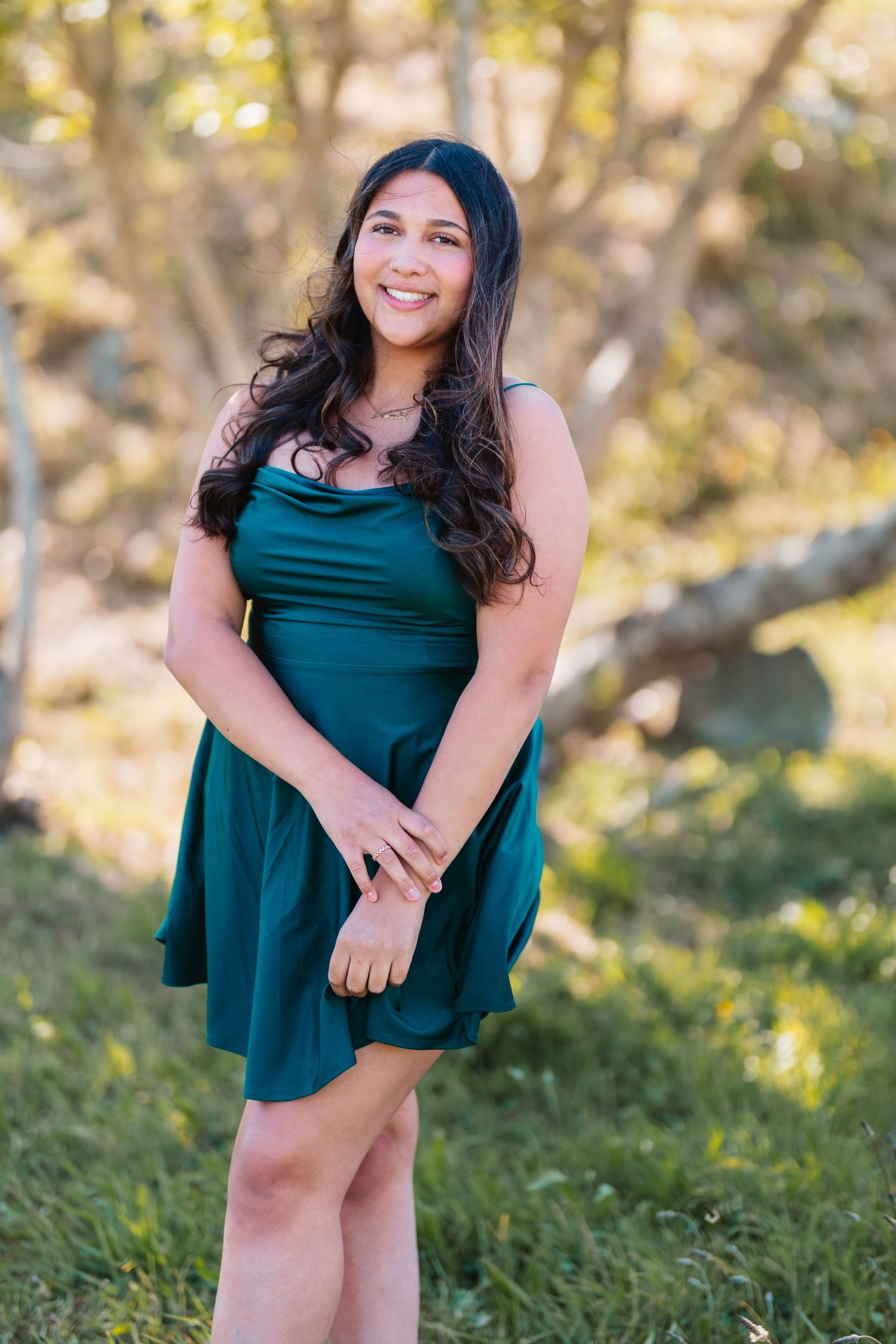 Young woman in teal dress smiling outdoors among trees and green grass in natural light portrait.