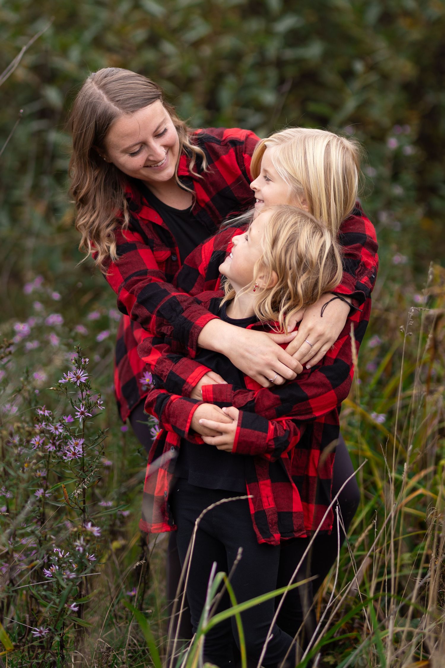 A mother hugs her two blonde daughters in a field, all wearing matching red plaid flannel shirts.
