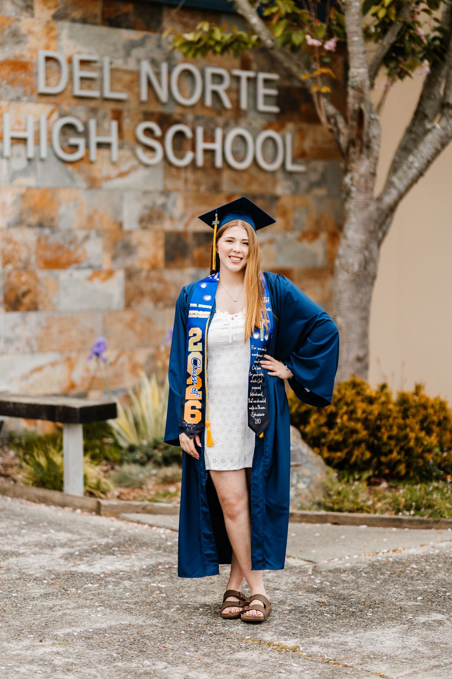 Smiling graduate in blue cap and gown with 2025 sash poses in front of Del Norte High School sign.