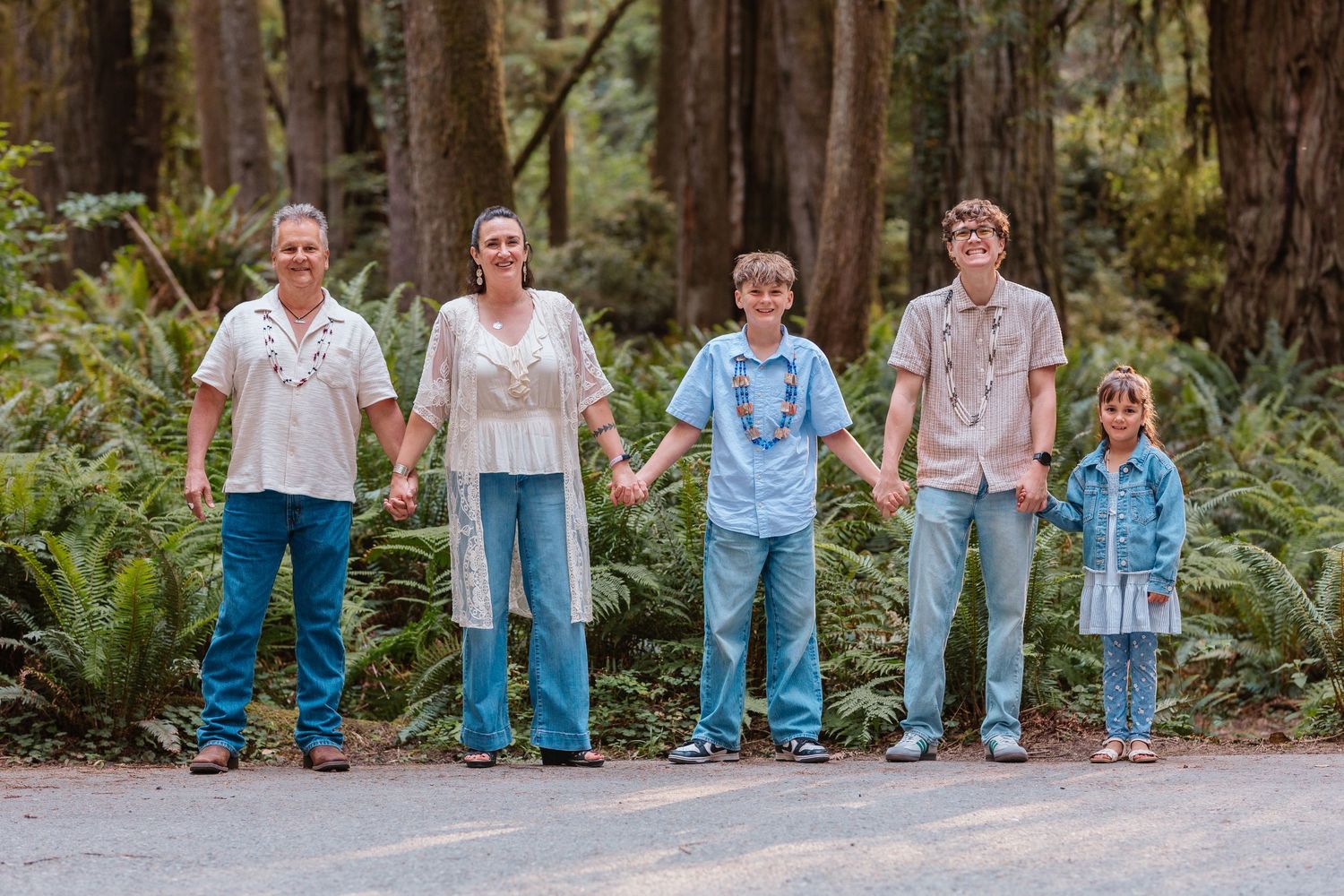 Family of five holding hands on a forest path surrounded by lush green ferns and trees.