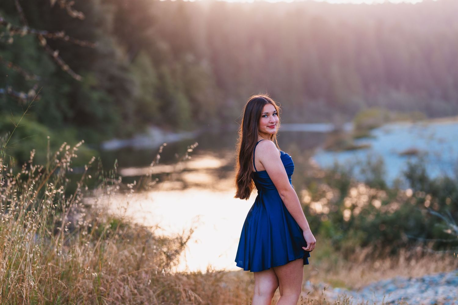 Teen girl in blue dress stands near a sunlit river surrounded by golden grass and pine trees.
