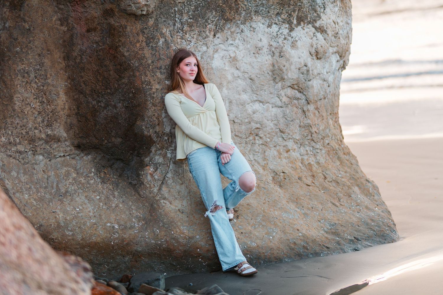 Young woman in light cardigan and jeans posing against a rocky cliff on the beach.