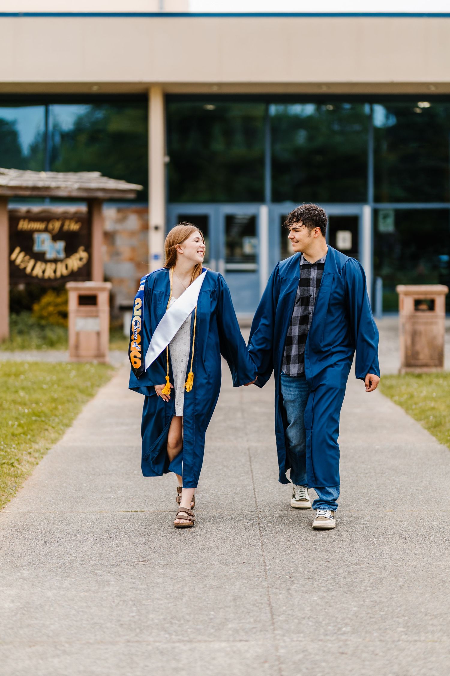 Two graduates in blue gowns walk hand in hand outside a school building labeled Home of the Warriors.