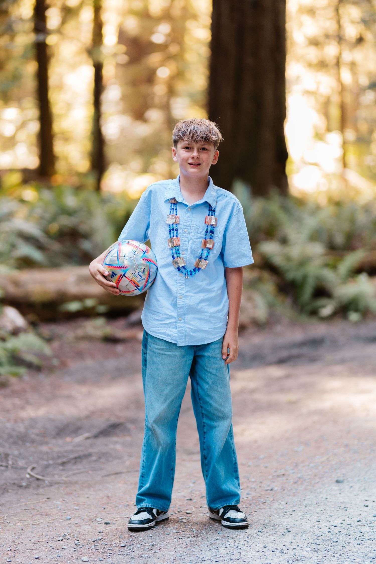 A boy standing on a forest trail holding a soccer ball, wearing a blue shirt, jeans, and a beaded necklace.