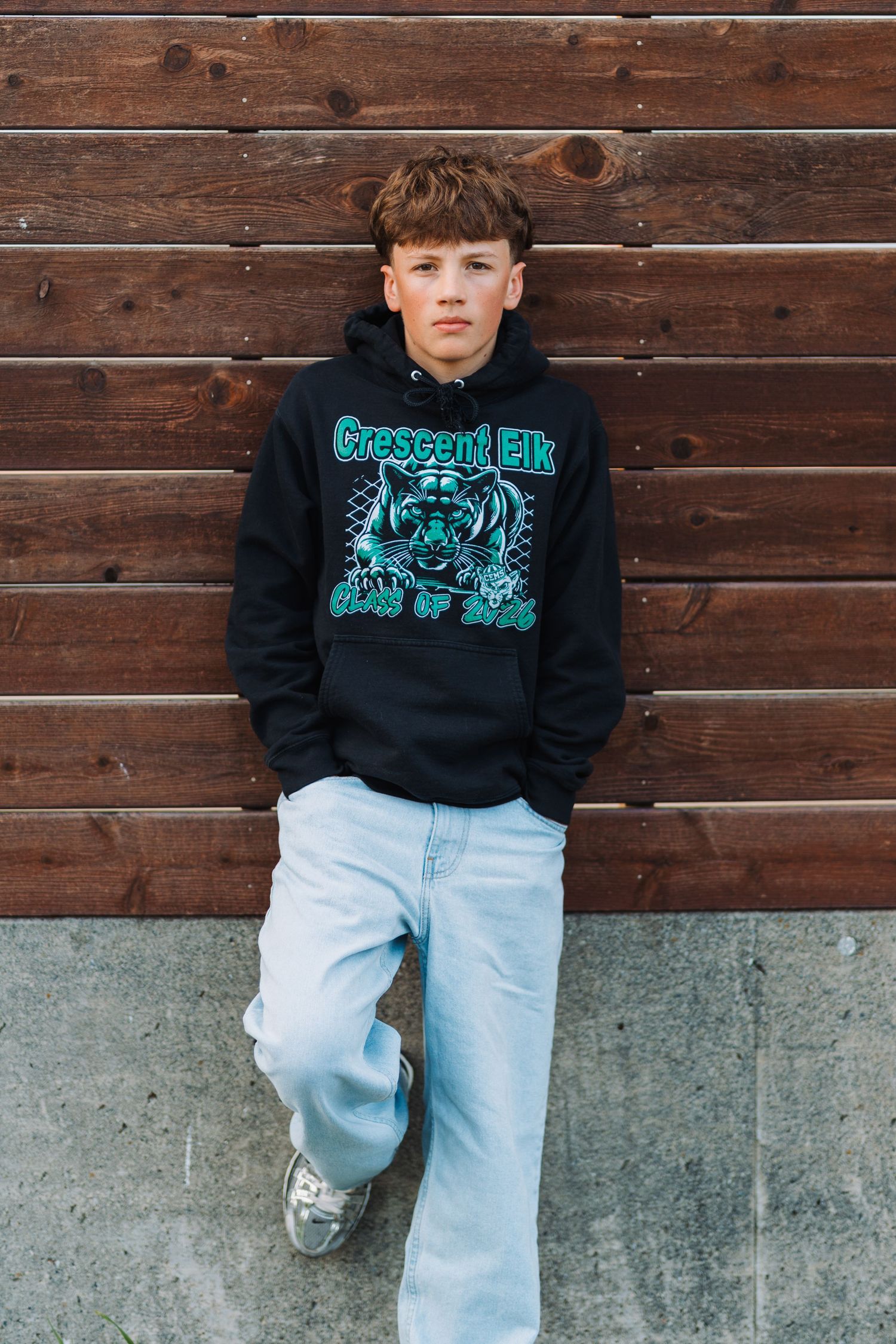 Teen boy in black Crescent Elk graphic hoodie and light wash jeans leaning against wooden wall.