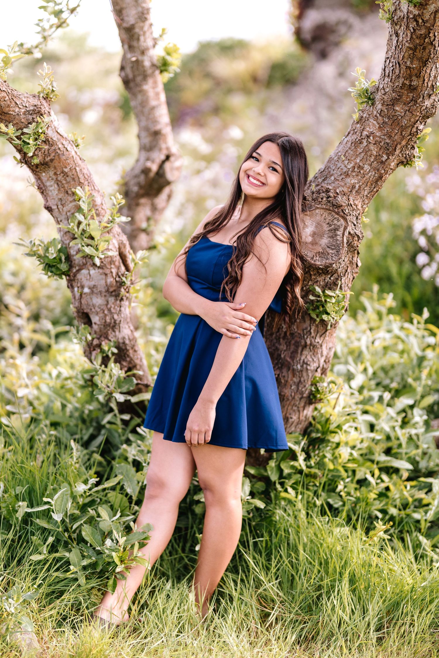 Young woman in navy blue dress smiling outdoors, leaning against a tree surrounded by lush green foliage.
