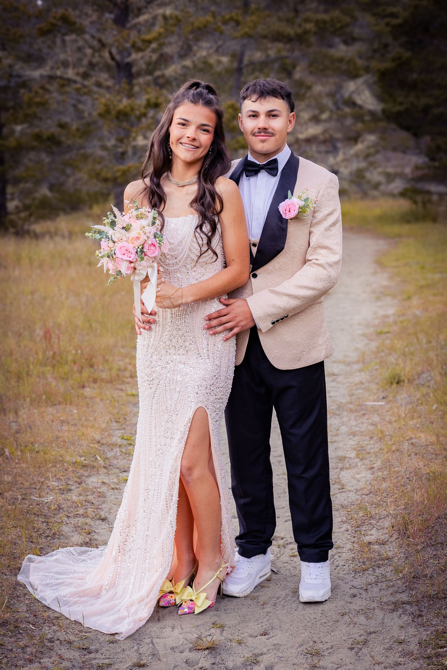 A young couple poses outdoors in prom attire, she in a beaded gown with bouquet, he in a tan tuxedo jacket.