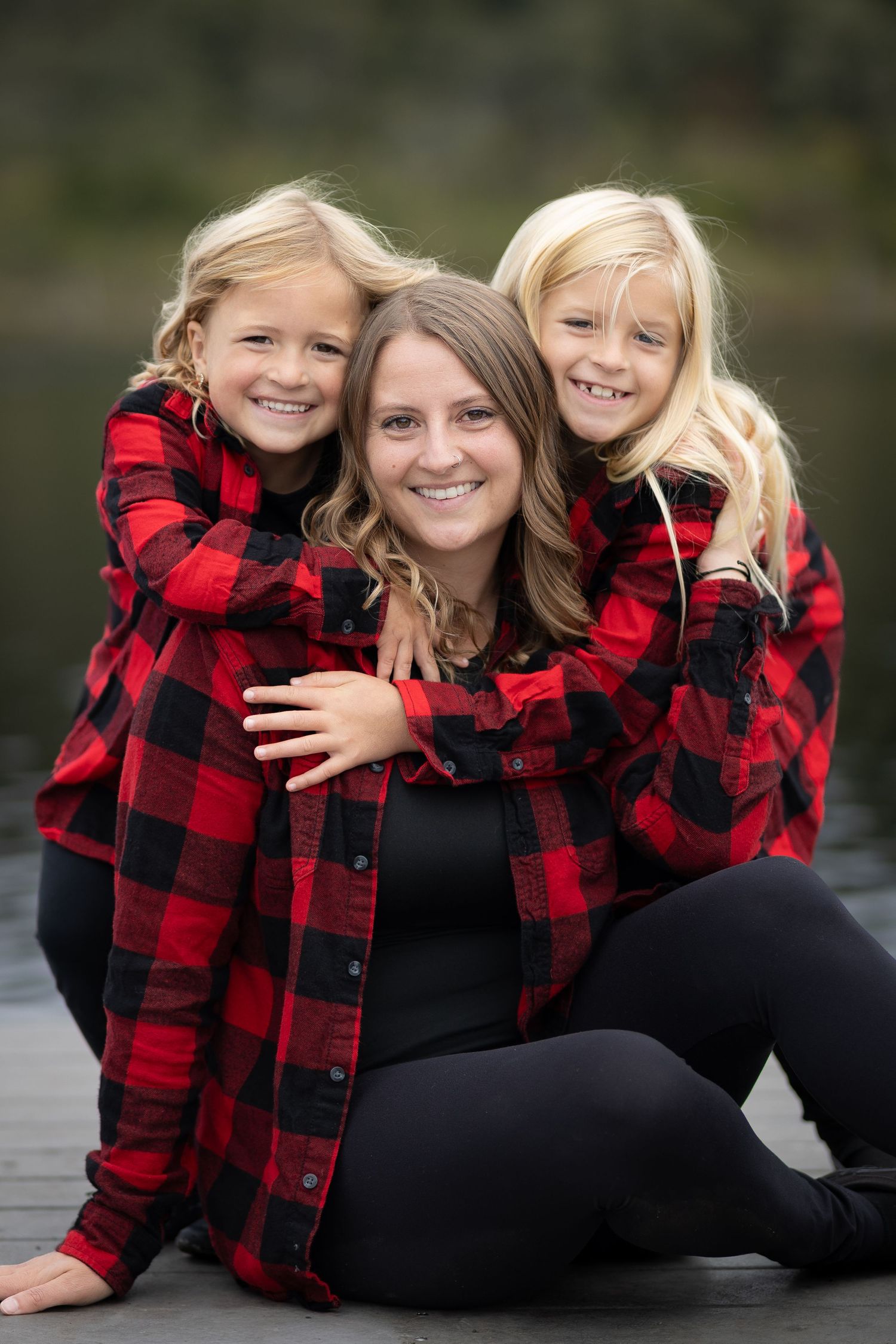 A happy mother and her two blonde daughters in matching red plaid shirts embrace on a dock.