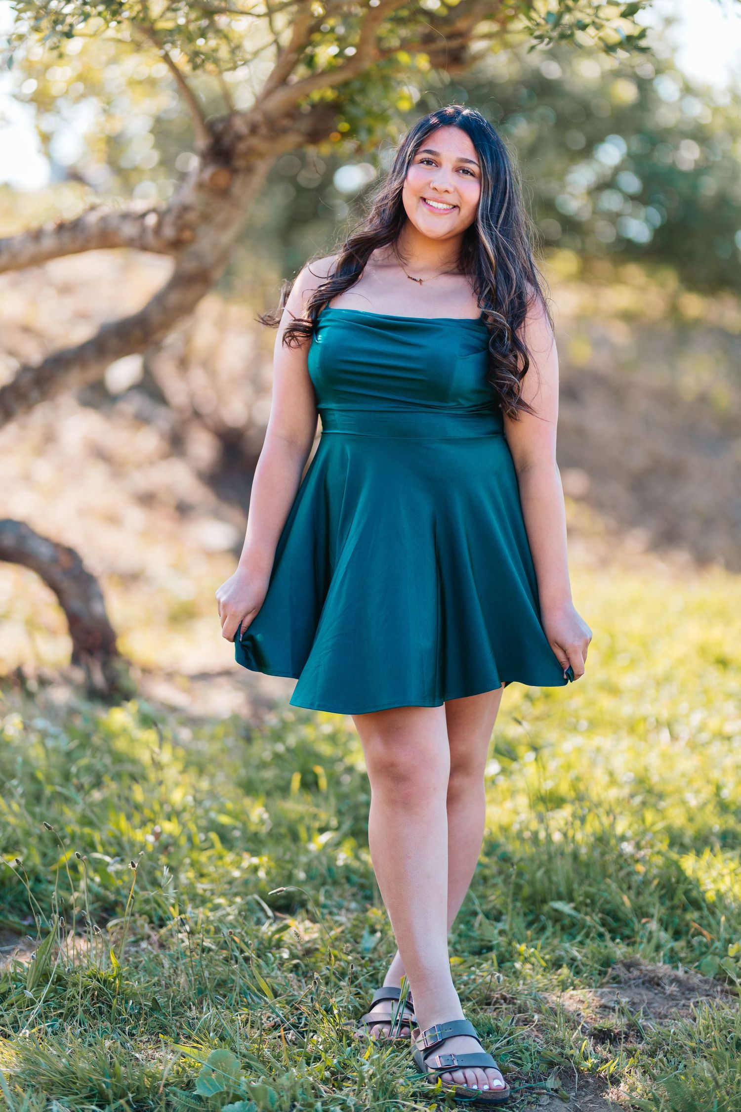 Smiling young woman in emerald green strapless skater dress standing on grass near a tree.