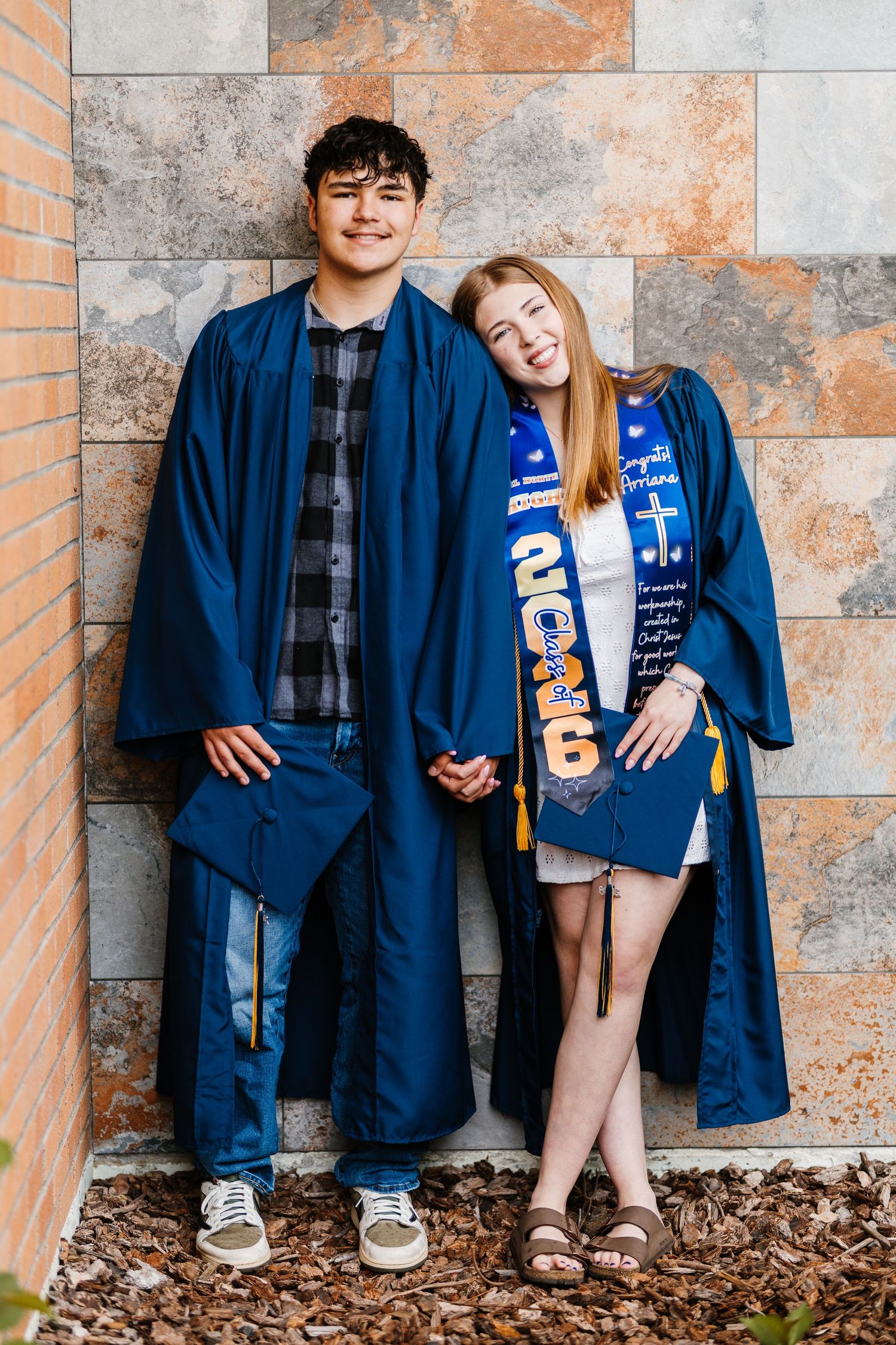 Two smiling graduates in blue caps and gowns pose together against a stone wall for their graduation photos.