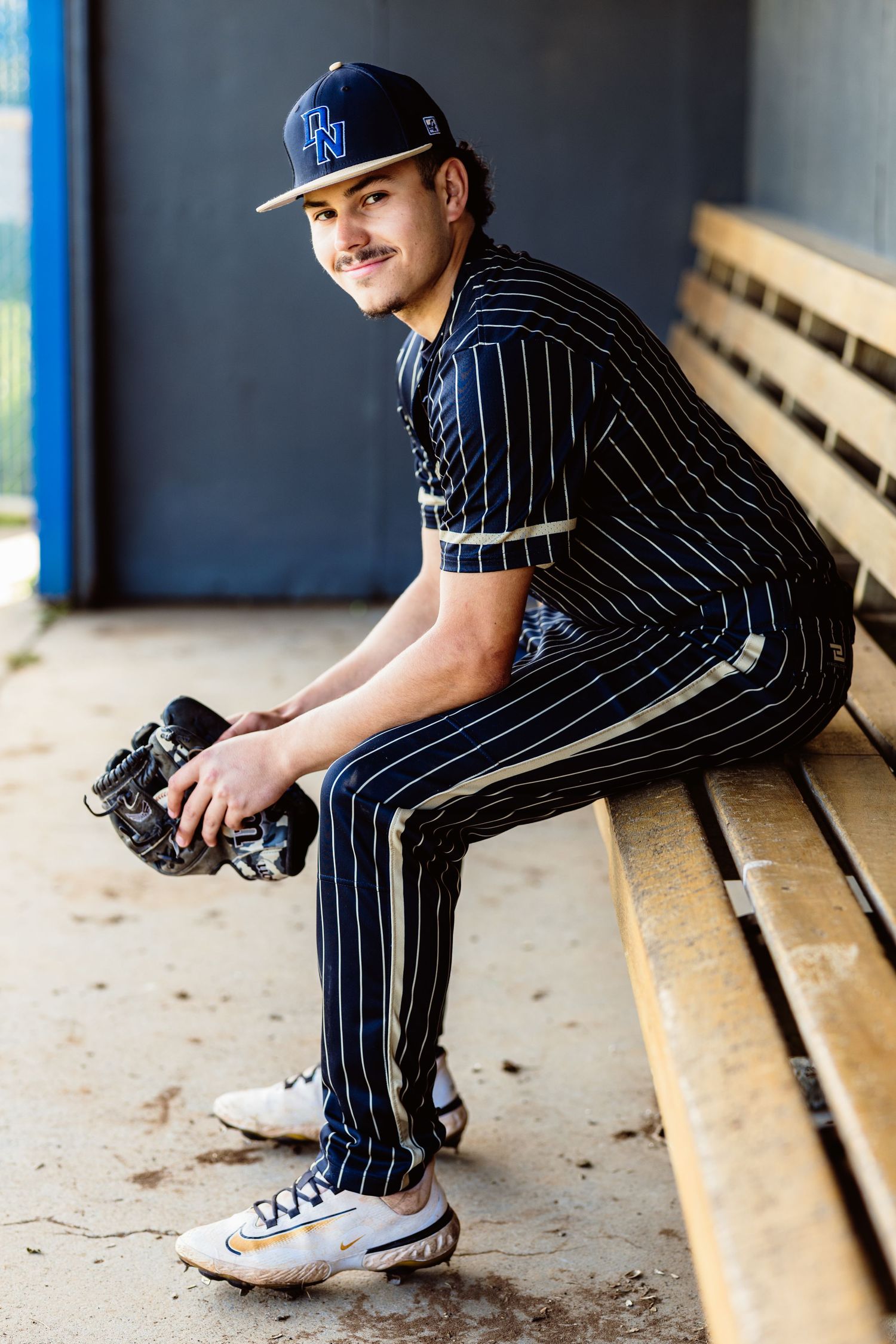 High school baseball player in pinstripe uniform sitting in dugout holding glove during senior portrait session.