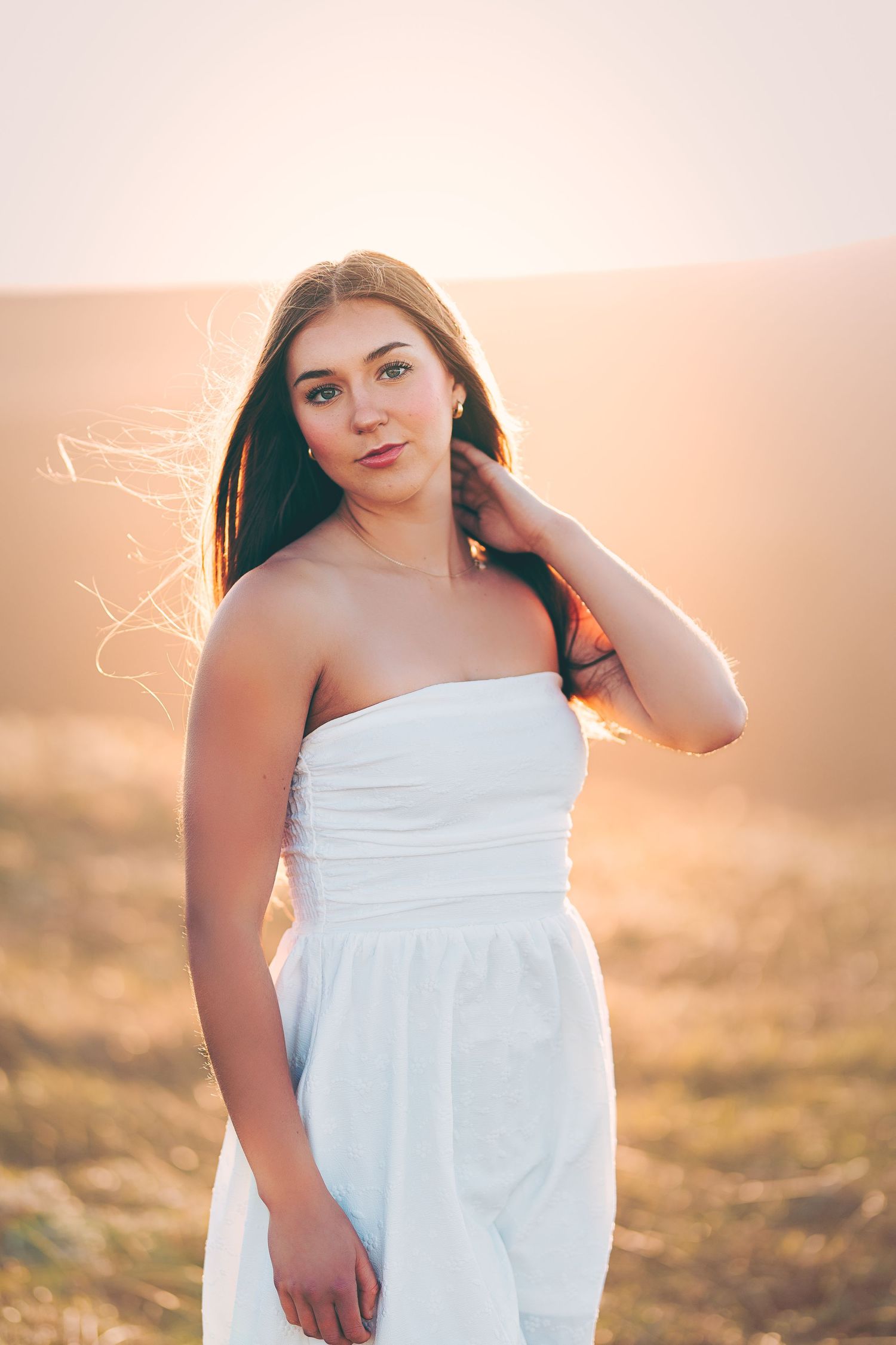 Young woman in white strapless dress poses in golden field at sunset with hair blowing in breeze.