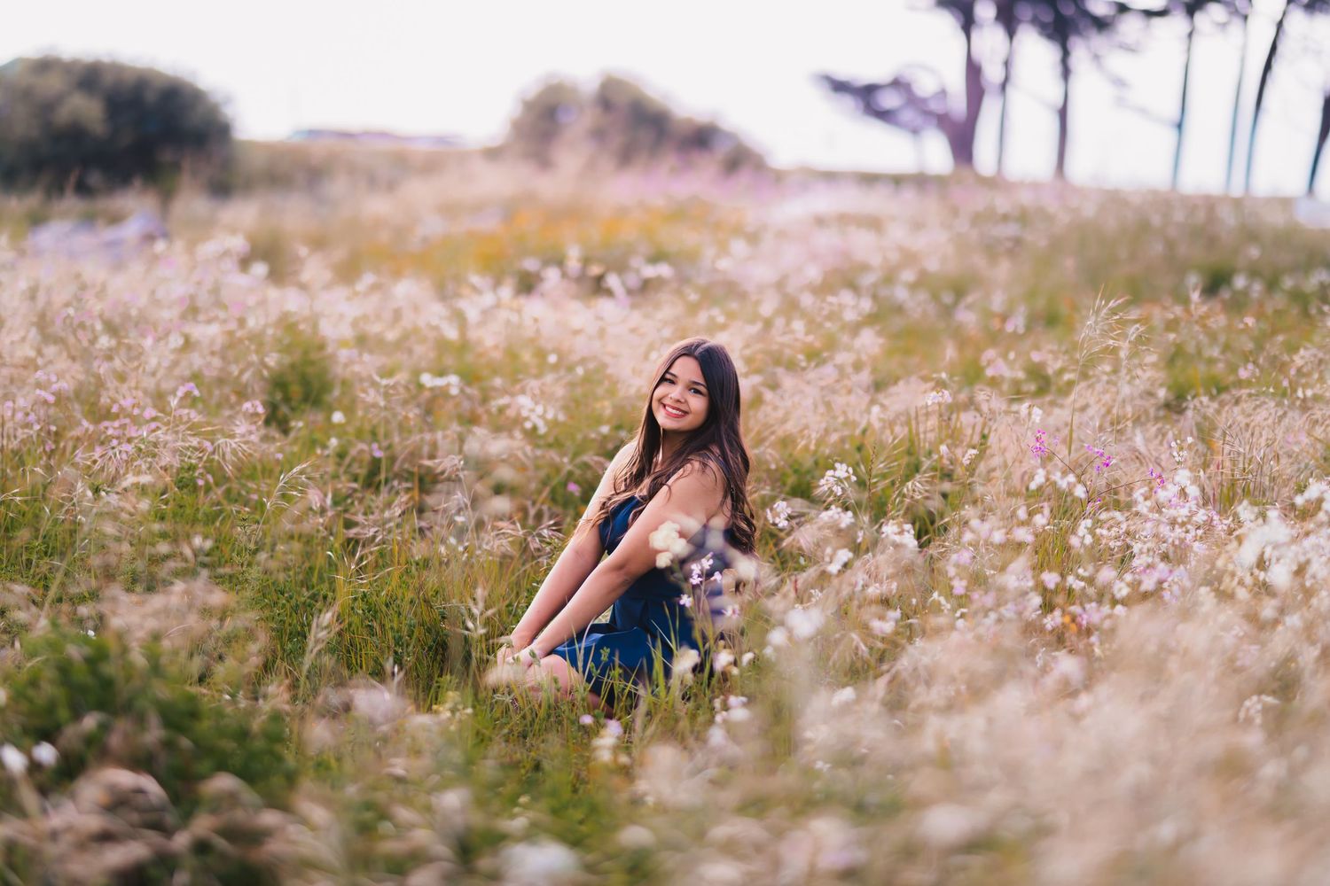 Smiling teenage girl sitting in a field of wildflowers wearing a blue dress during a senior portrait session.
