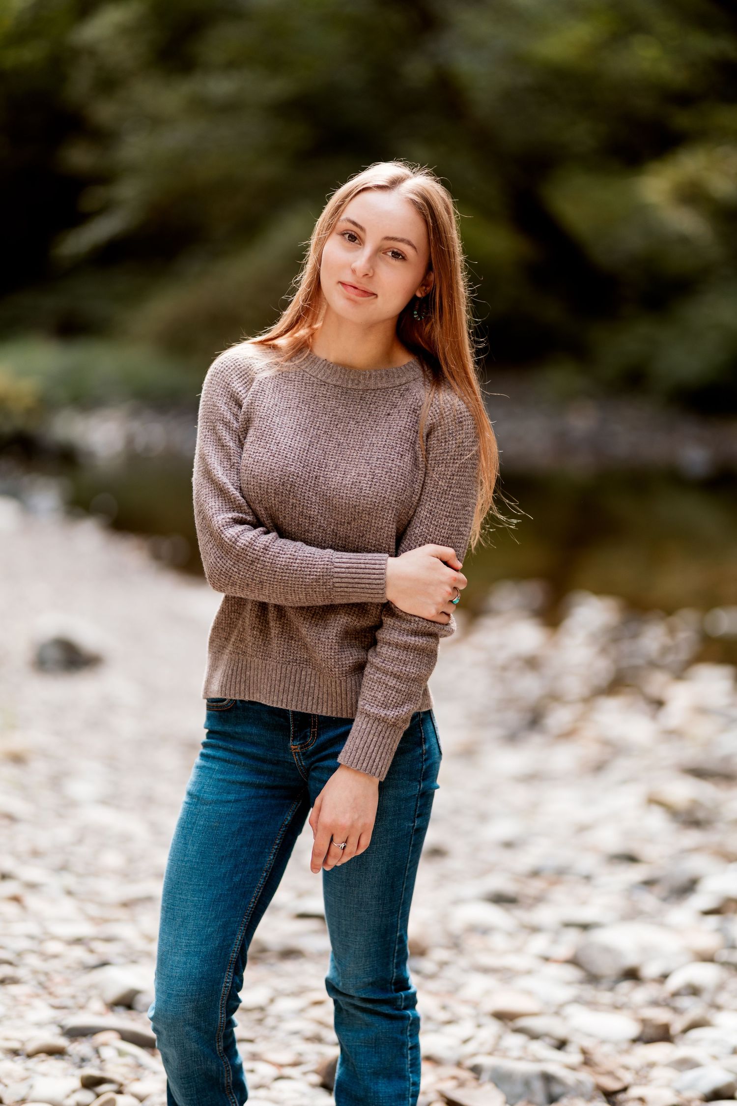 Young woman with long hair in brown sweater and jeans standing on rocky riverbank in sunlight.