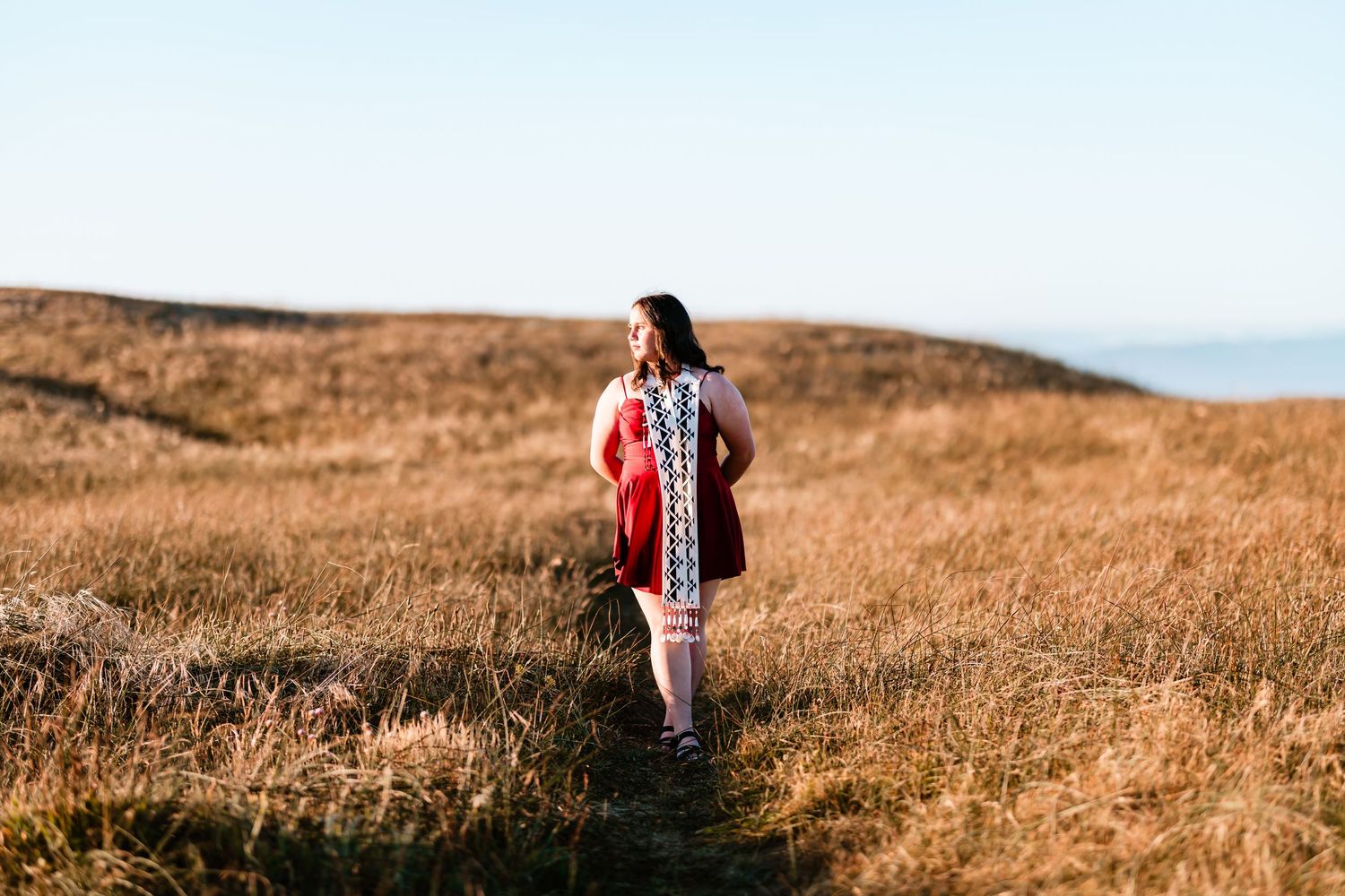 Woman in embroidered dress walking through golden meadow with ocean horizon in warm sunlight.