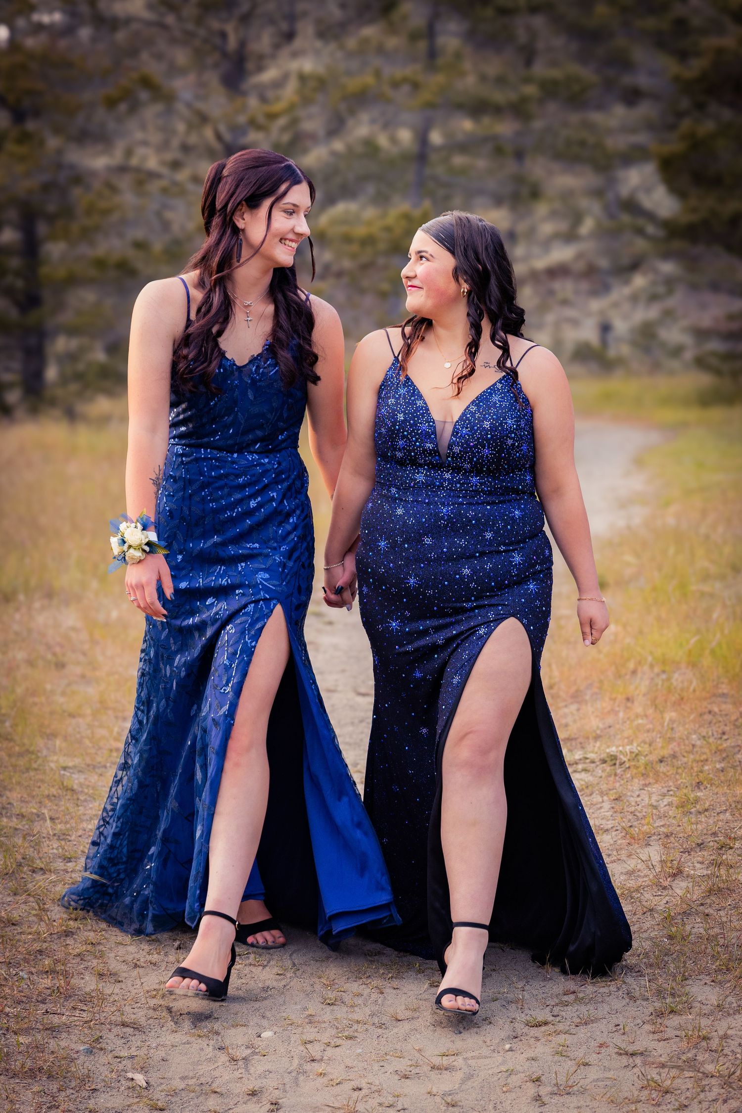 Two young women in matching blue sequin prom dresses with slits walk hand in hand on a dirt path.