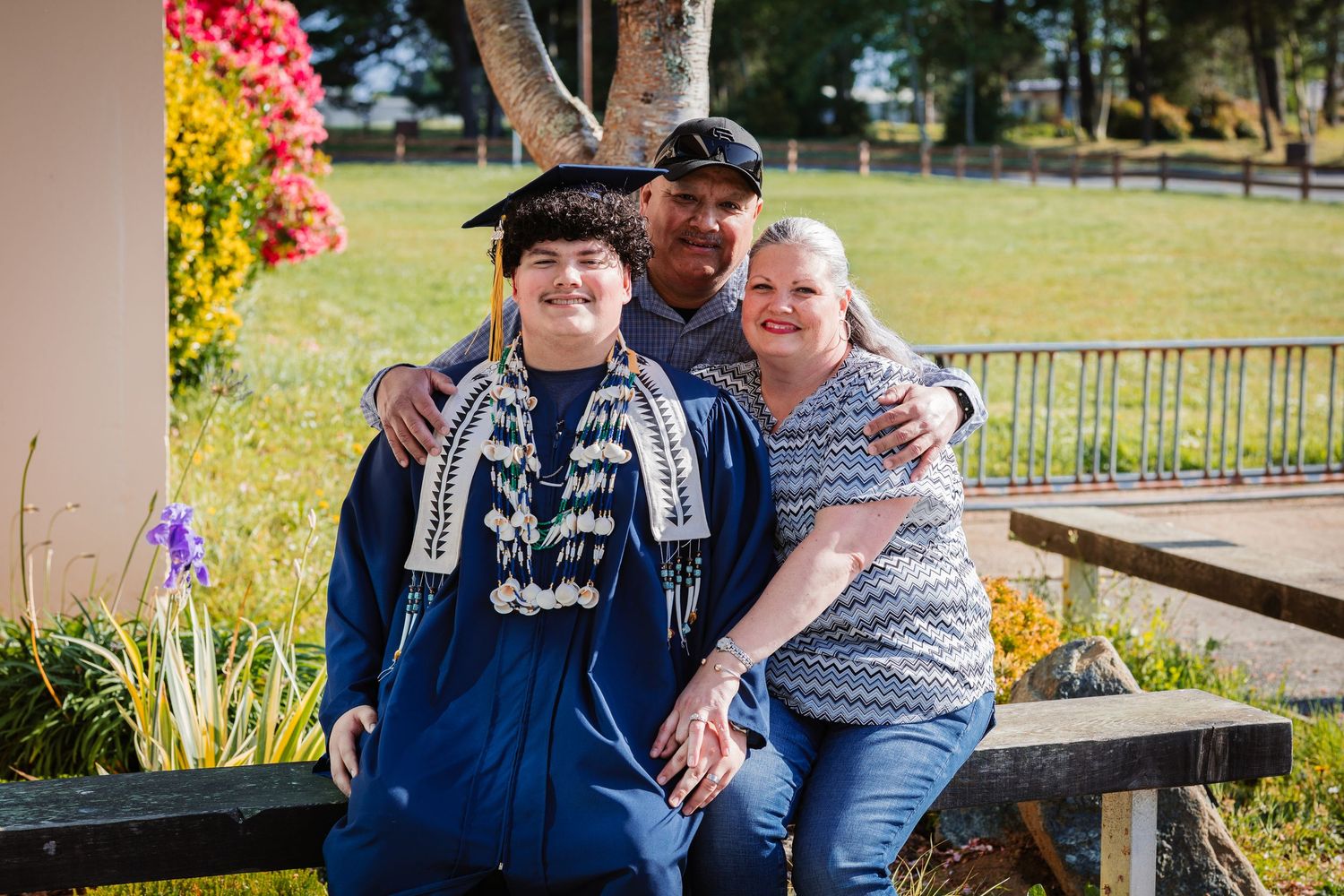 Graduate in blue gown and cap poses with two proud family members outdoors on a sunny day.