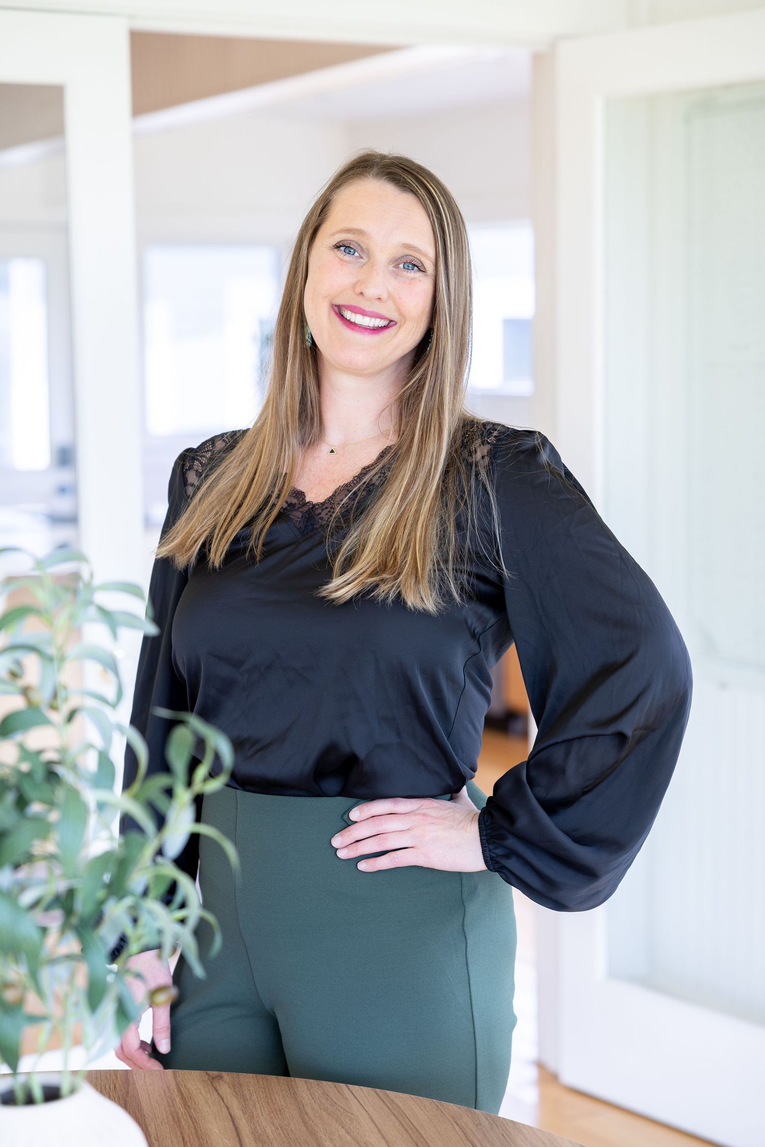 Professional woman in black blouse and green skirt smiling confidently in a bright modern interior setting.
