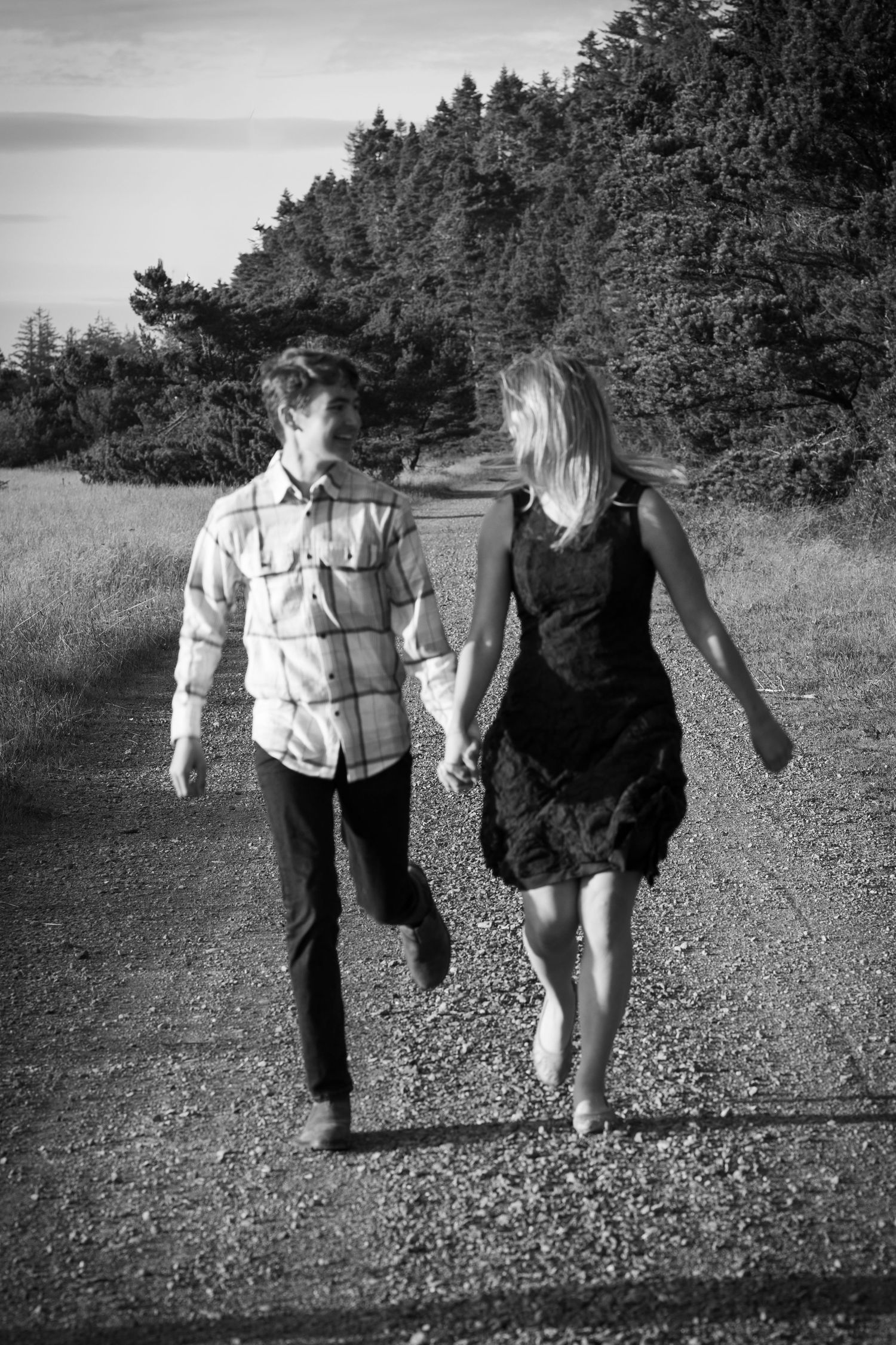 Black and white photo of a couple holding hands walking down a gravel path surrounded by trees.