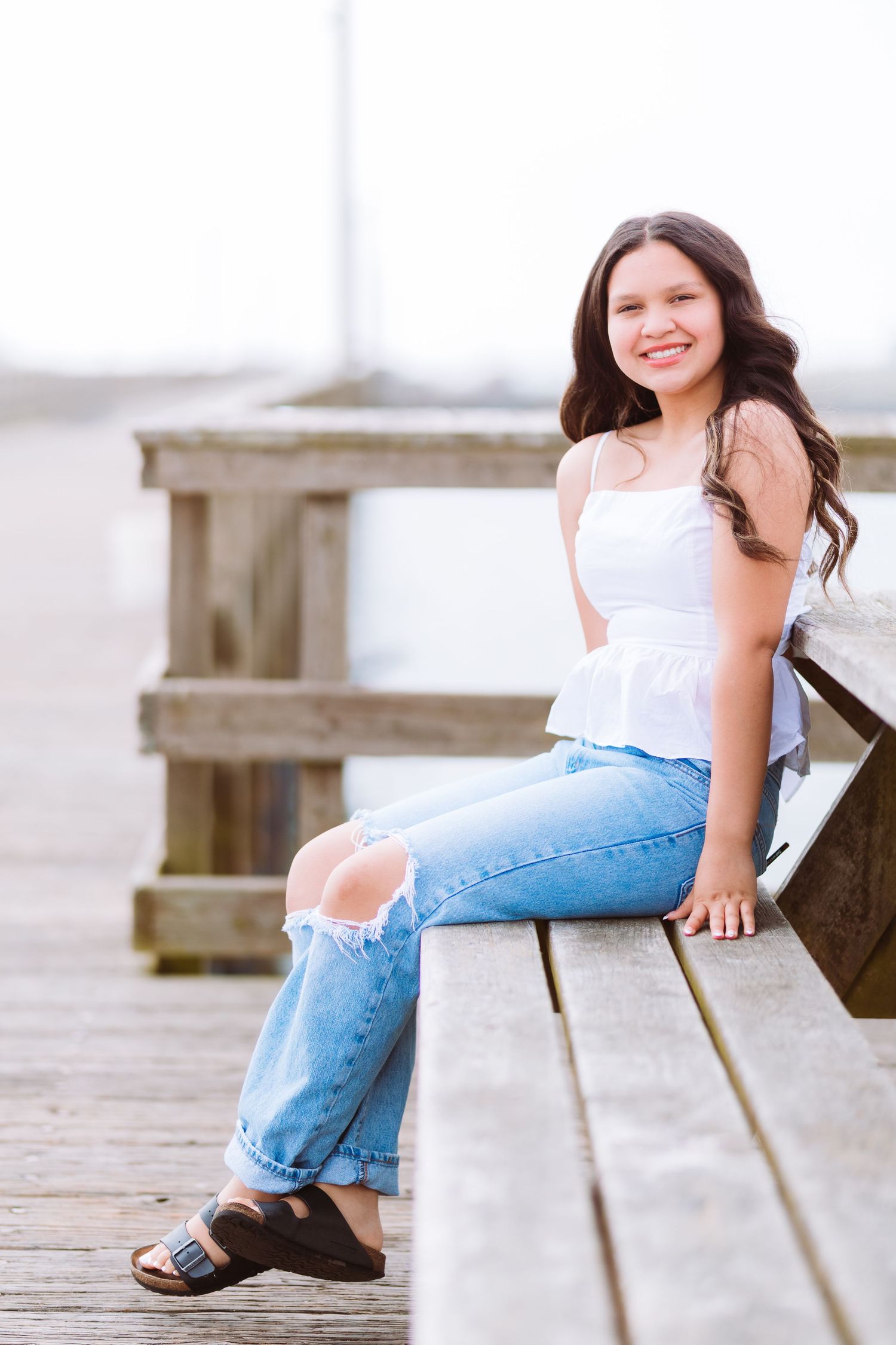 Young woman in white top and ripped jeans sitting on wooden pier bench, smiling outdoors near waterfront.