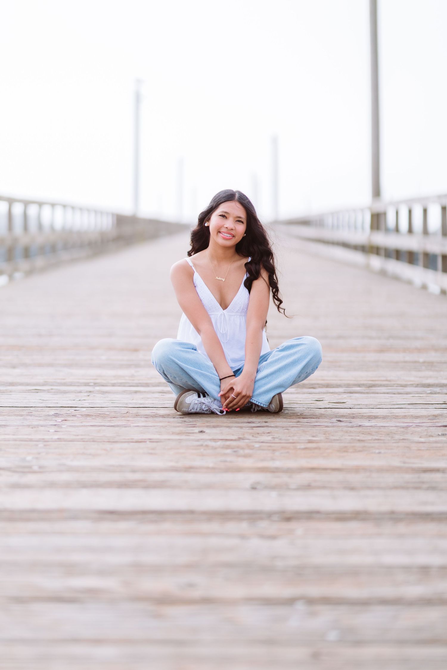 Young woman with long dark hair sits cross-legged on a wooden pier, smiling, wearing white top and light blue jeans.