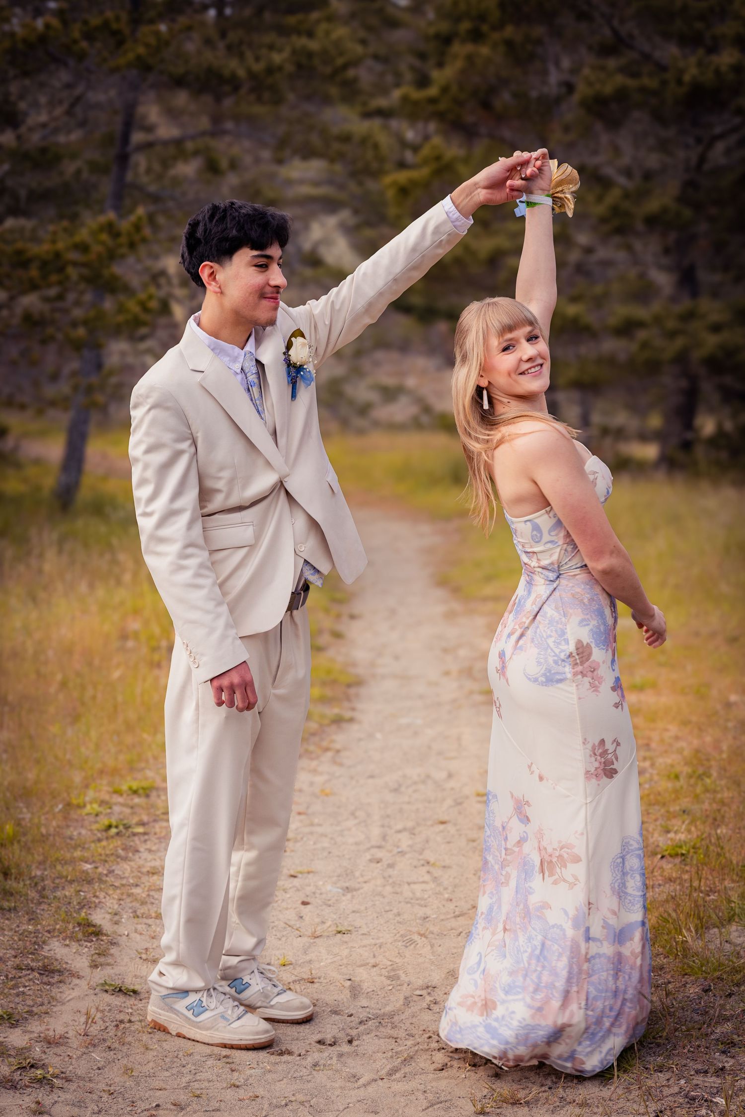 Smiling prom couple dance outdoors, boy in tan suit twirls girl in flowing floral dress on dirt path.
