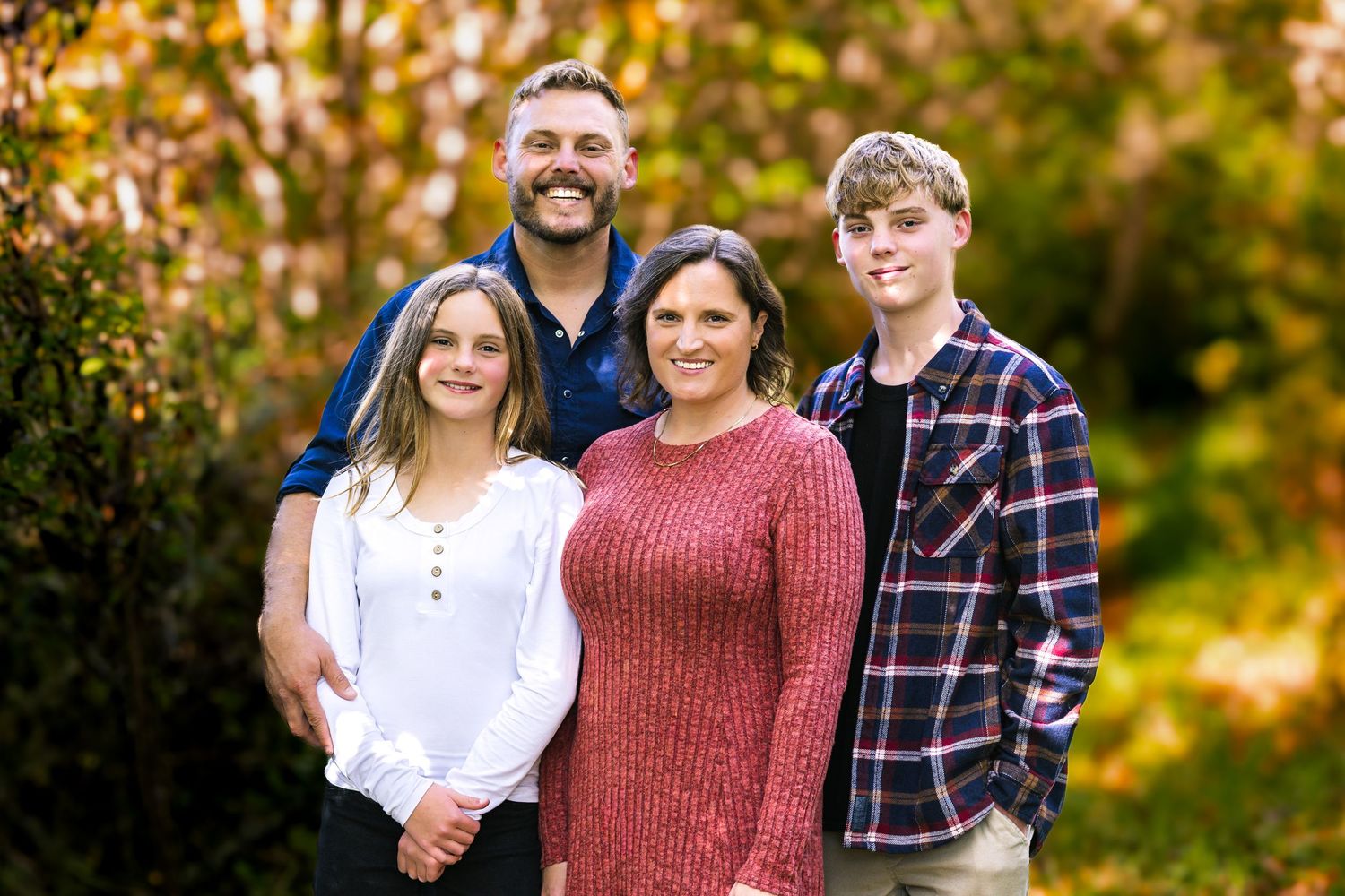 Family of four smiling outdoors in autumn, posing together among colorful fall foliage for a portrait.