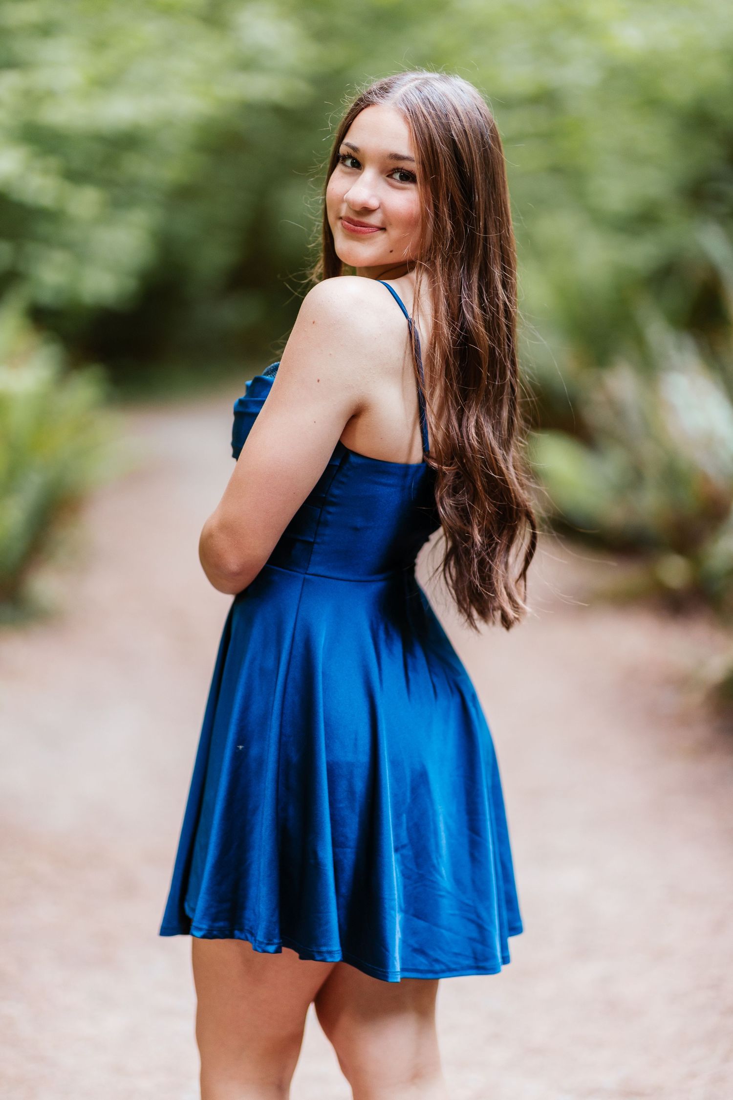 Teen girl in royal blue satin dress poses on garden path, long brown hair, smiling over shoulder.