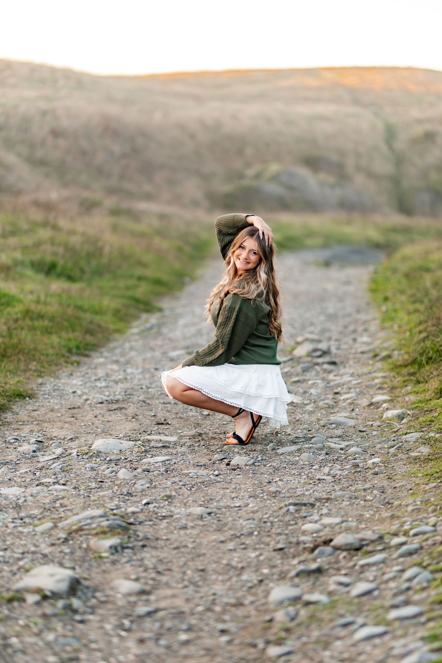 Young woman in green sweater and white skirt crouching on a rocky gravel path outdoors.