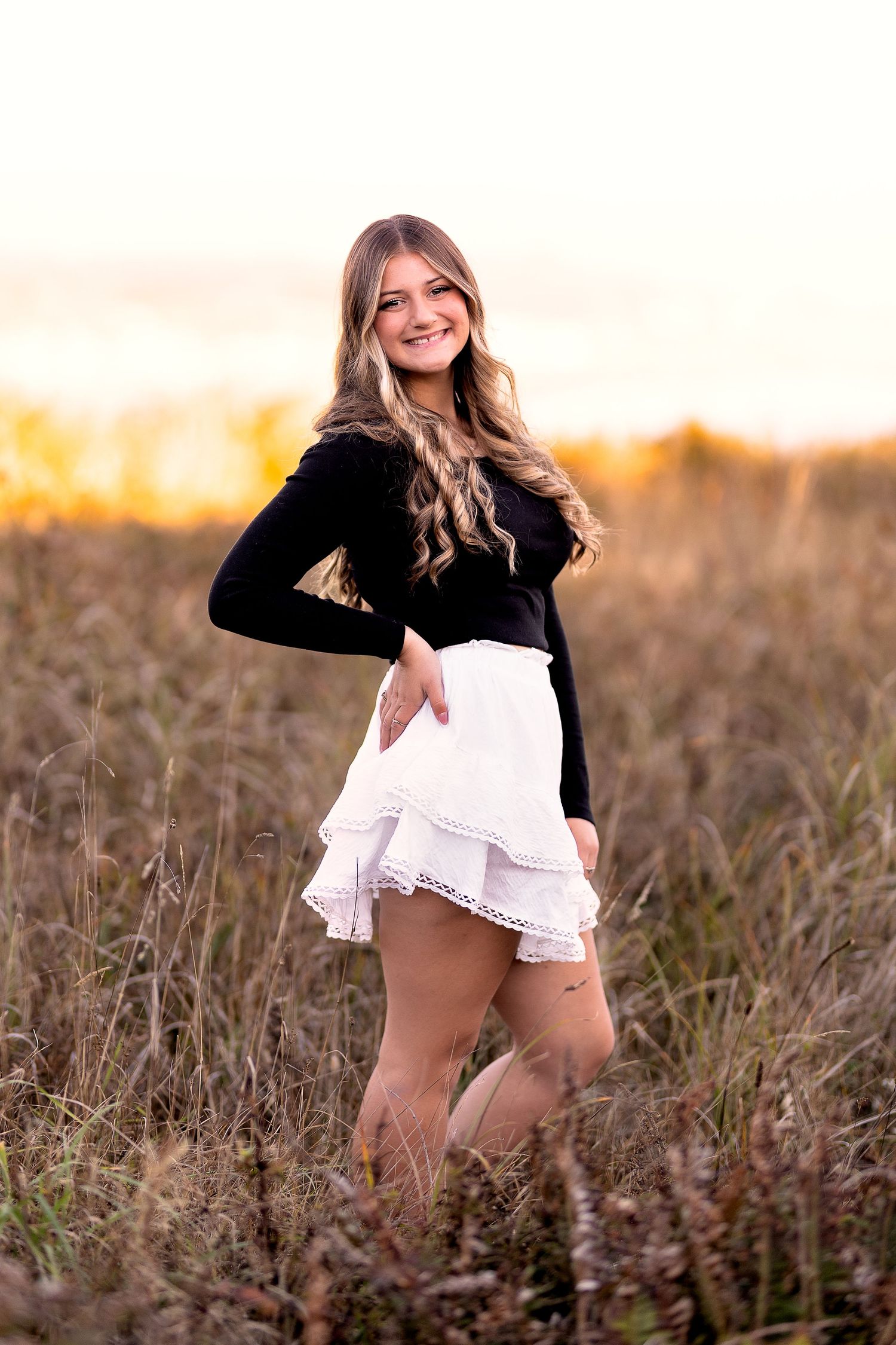 Blonde teenage girl in black top and white ruffled skirt poses in golden field at sunset.