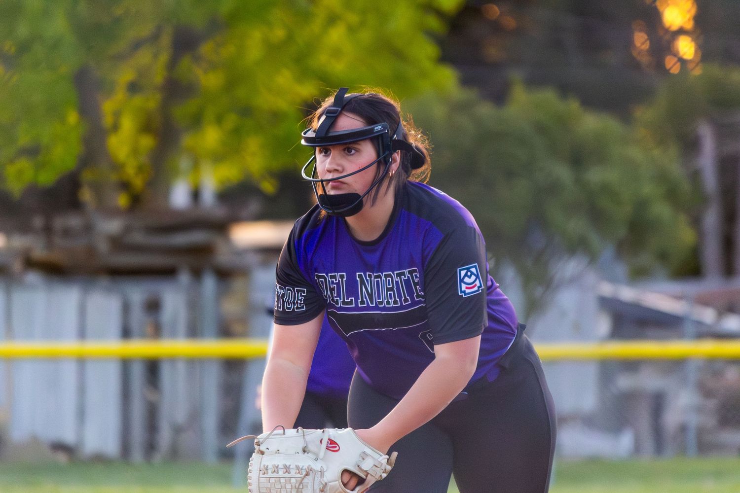 Female softball player in purple uniform stands ready in fielding position during an outdoor game.