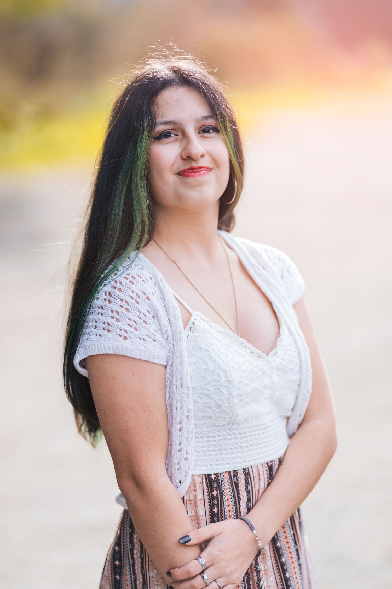 Young woman with green-highlighted dark hair smiles outdoors wearing a white top and striped skirt.