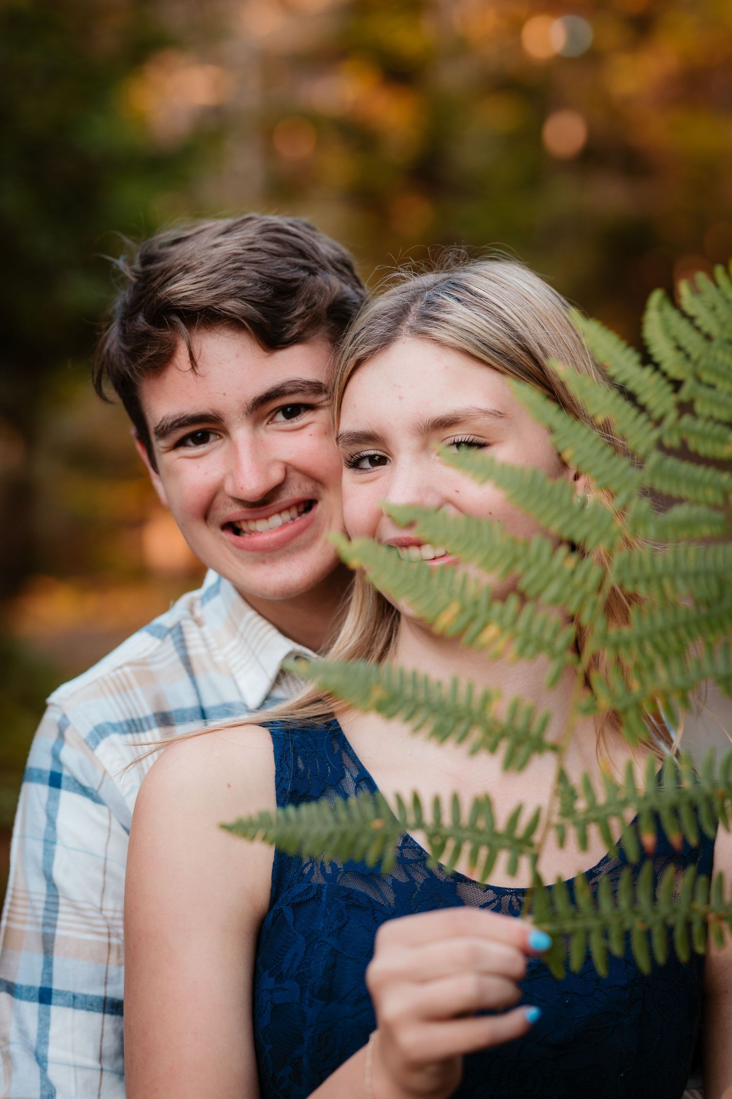 Young couple smiling behind green fern leaf in colorful autumn forest setting.