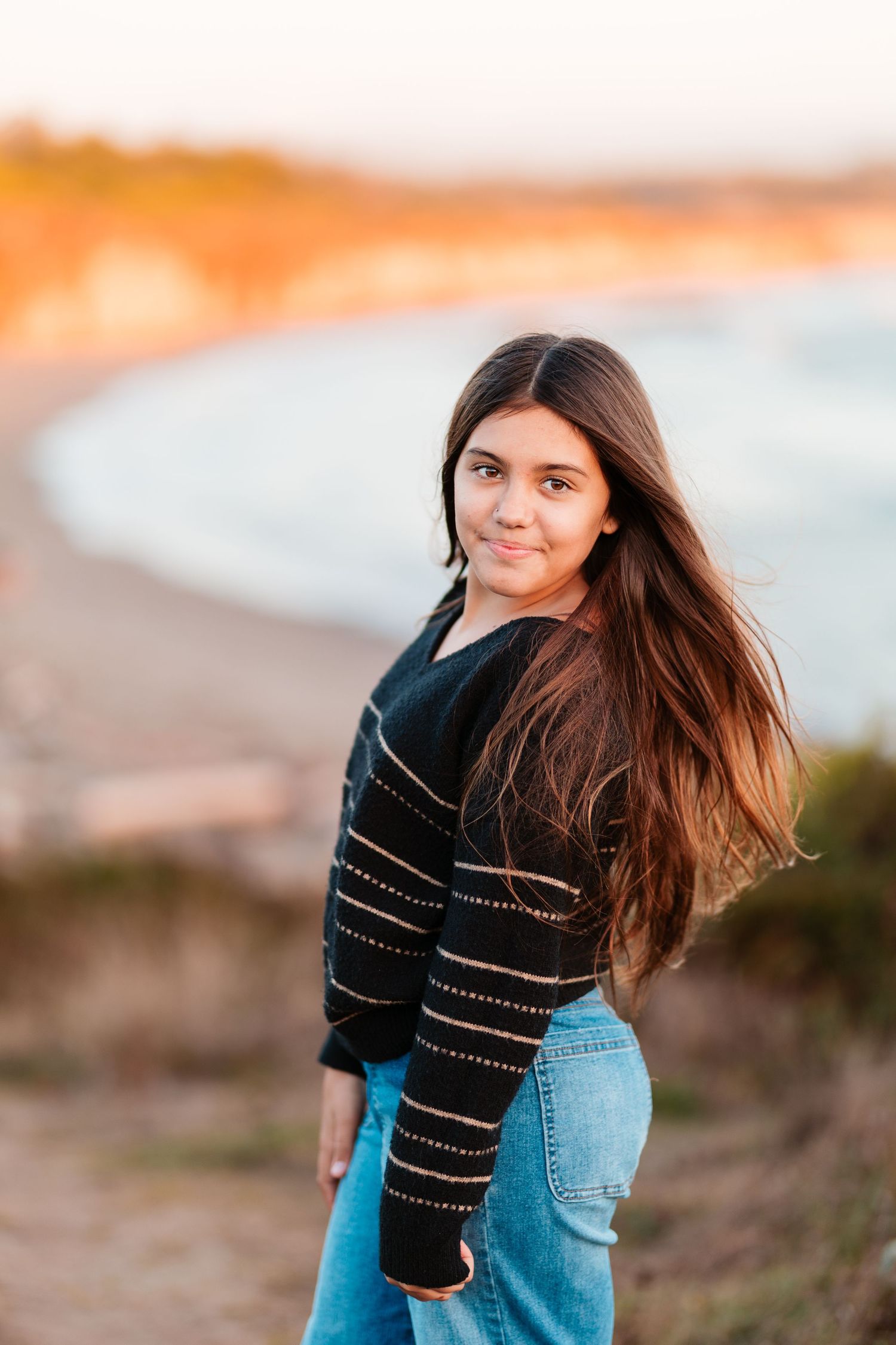 Young girl with long dark hair in black sweater and jeans posing near a scenic beach at sunset.