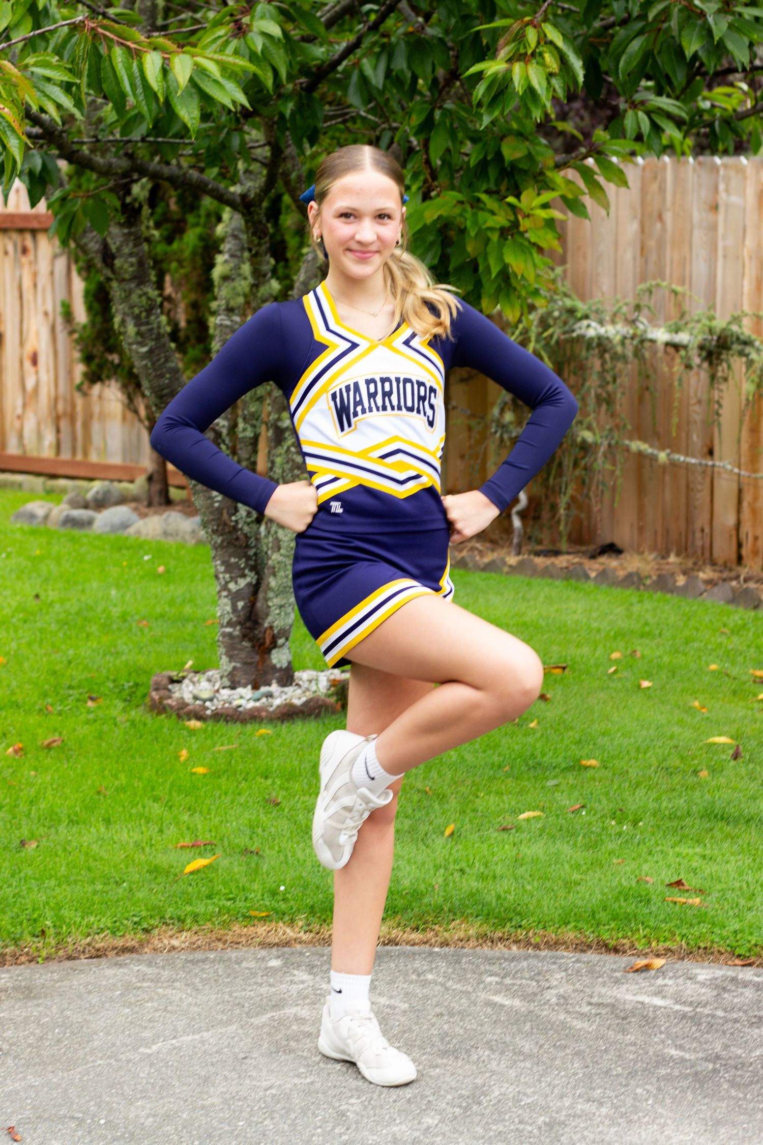 Teen girl in Warriors cheerleader uniform posing with one leg raised in backyard setting.