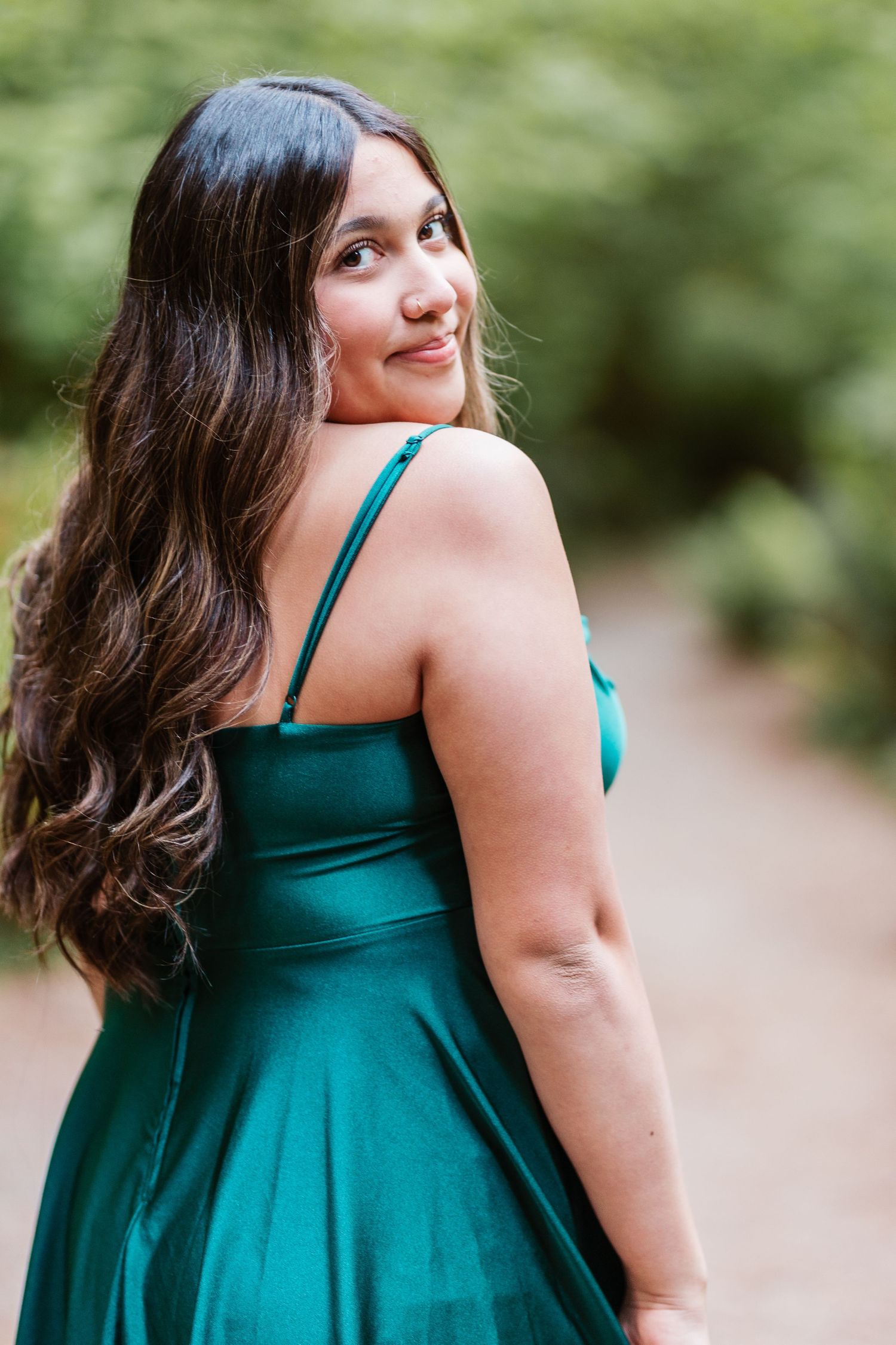 Young woman in emerald green satin dress with spaghetti straps, looking over shoulder, long wavy hair, outdoor path.
