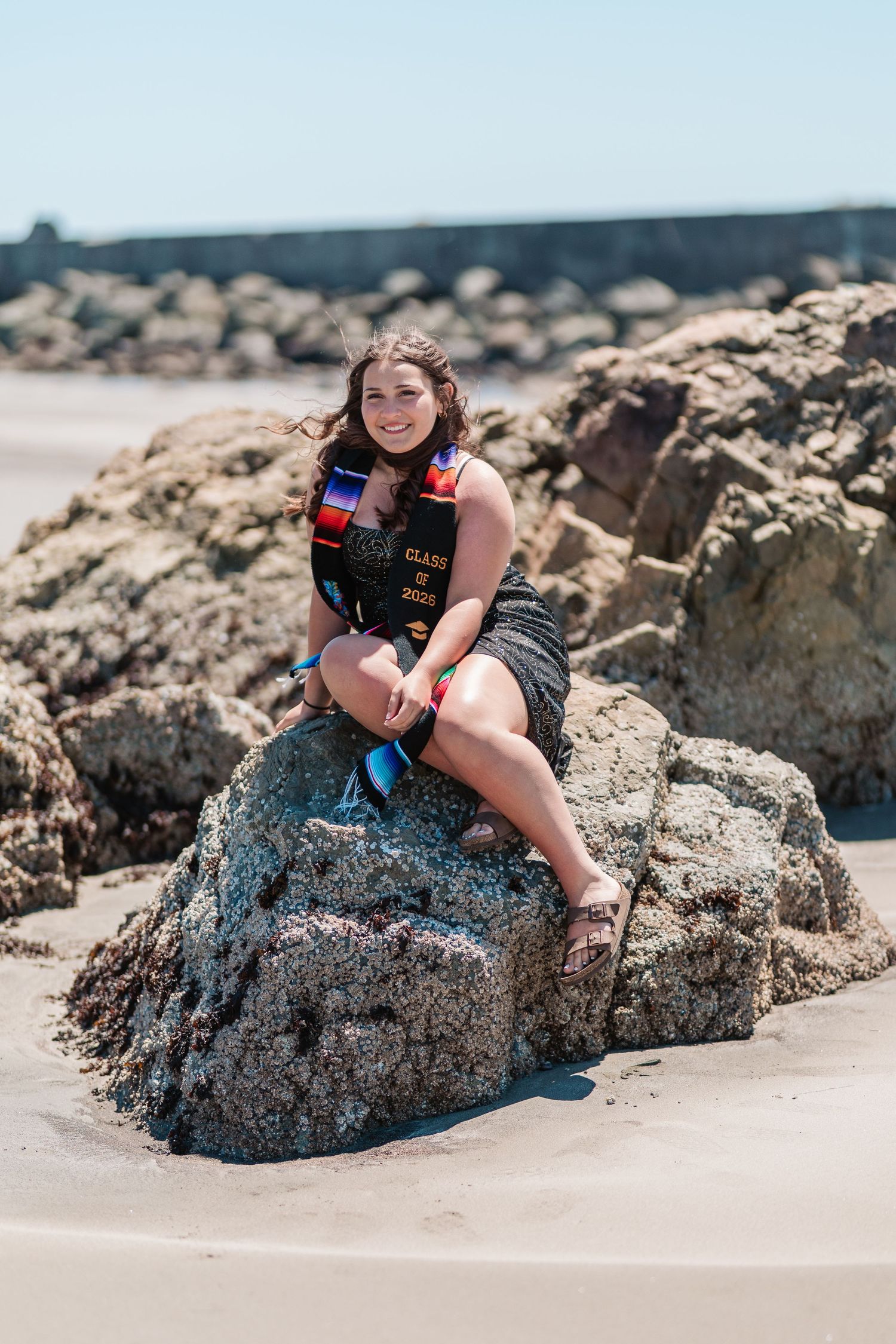 Smiling graduate in cap and gown sitting on beach rocks, celebrating Class of 2023 on a sunny day.
