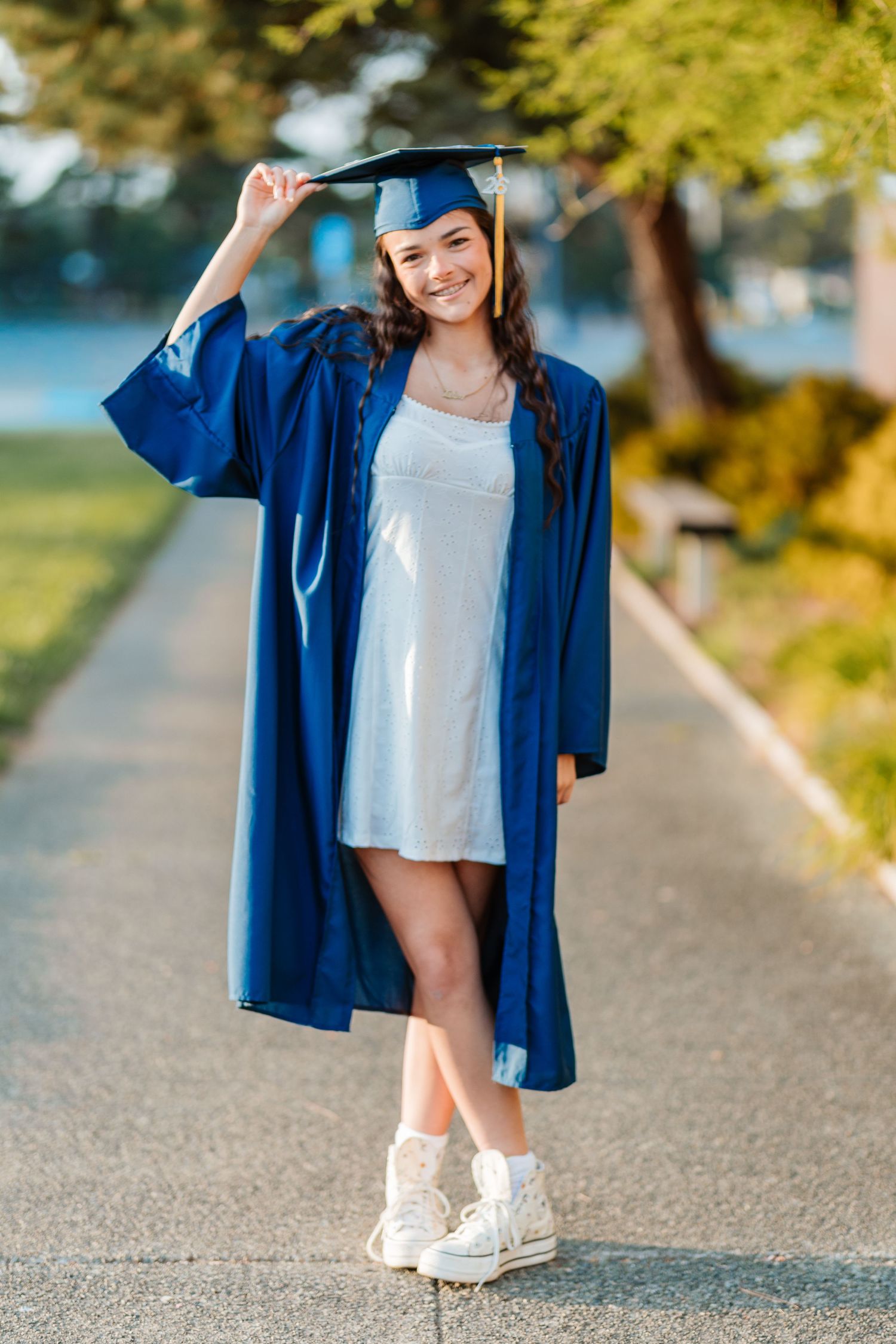 Smiling graduate in blue cap and gown poses outdoors in white dress and sneakers on a sunny day.